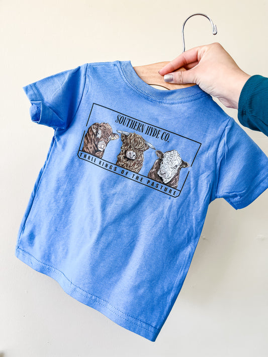 A hand holds the light blue Pasture Kings T-Shirt by Southern Hyde Co, featuring illustrations of three cows with "Southern Hyde Co." above and "The Kings of the Pasture" below the artwork.