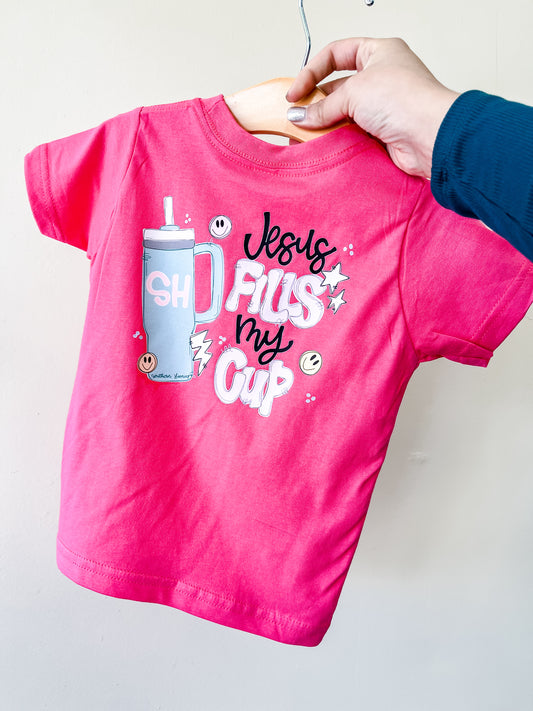 A hand holds up the Southern Hyde Co Jesus Fills My Cup T-Shirt, a bright pink toddler shirt featuring a tumbler cup graphic and playful “Jesus fills my cup” white lettering with stars and smiley faces.