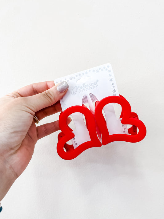 A hand with silver nail polish holds a pack of two red Heart Hair Clips from love & repeat, attached to a white card against a plain white background.