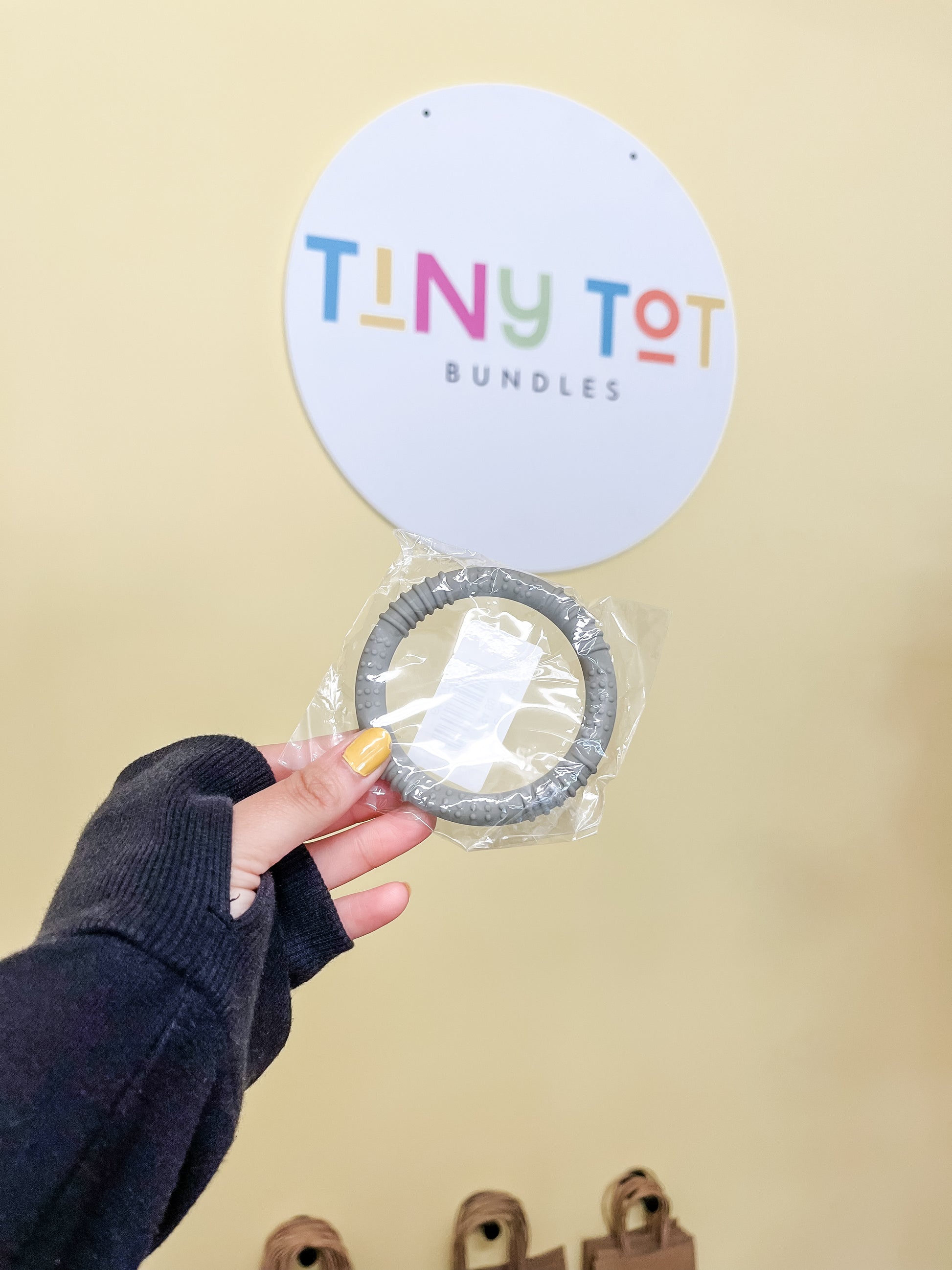 A hand with yellow nail polish holds a gray Circle teether by Tiny Tot Bundles in a plastic wrapper. In the background, a round sign with the brand name in colorful letters hangs on a beige wall.