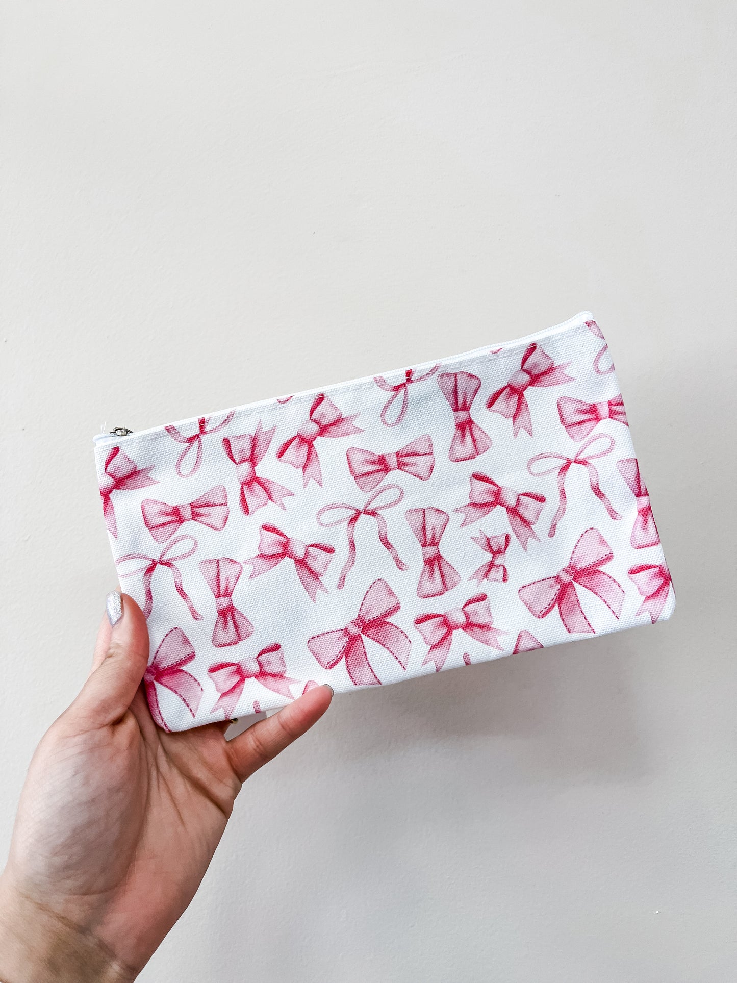 A hand holds the love & repeat Pretty in Bows Zipper Pouch, a white pouch featuring a pink bow pattern, against a plain light background.