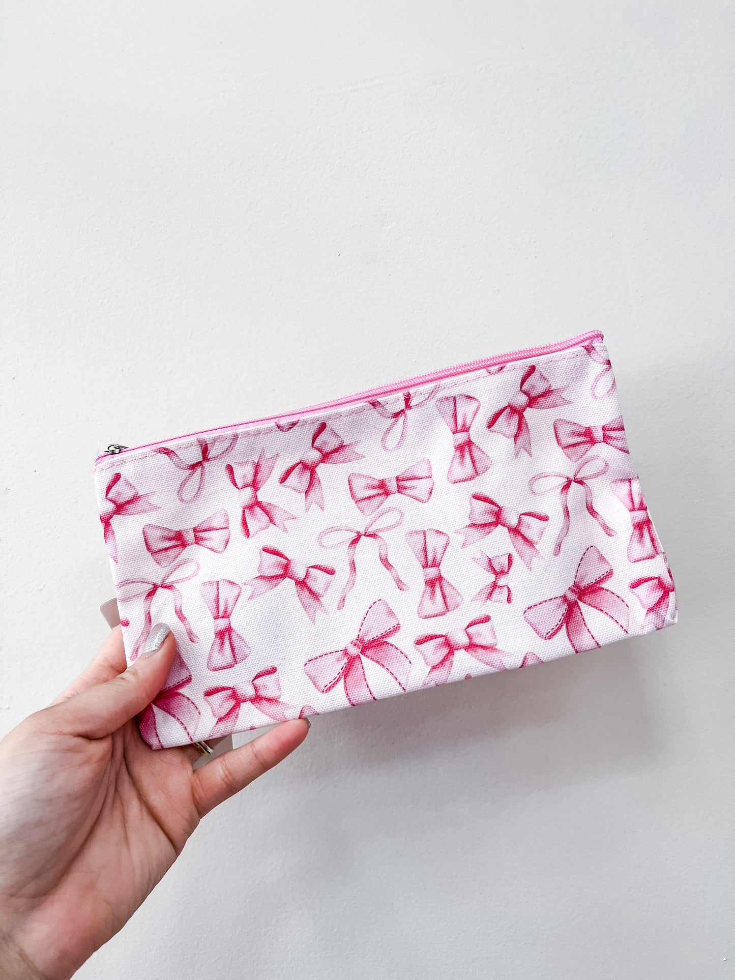 A hand holds the "Pretty in Bows Zipper Pouch" by love & repeat, a white pouch adorned with a pink bow pattern, set against a simple light background.