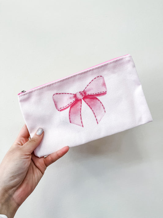 A hand holds the love & repeat Pretty in Bows Zipper Pouch, featuring a light pink color and a stitched pink bow design, shown against a plain white background.