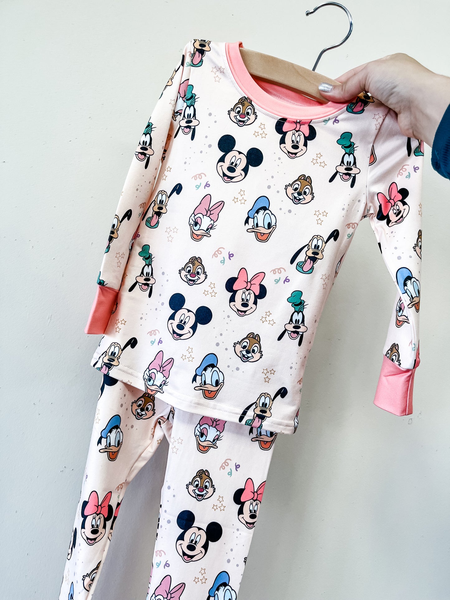 The Magic Friends Jammies Set by Tiny Tot Bundles is a two-piece children's pajama set in light pink with coral cuffs, featuring an all-over print of Disney characters like Mickey, Minnie, Donald, Daisy, Goofy, and Pluto.