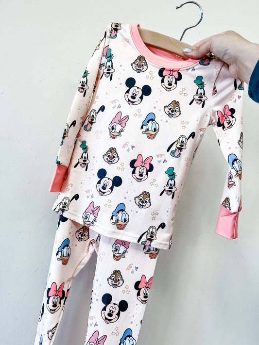 The Magic Friends Jammies Set by Tiny Tot Bundles is a two-piece children's pajama set in light pink with coral cuffs, featuring an all-over print of Disney characters like Mickey, Minnie, Donald, Daisy, Goofy, and Pluto.
