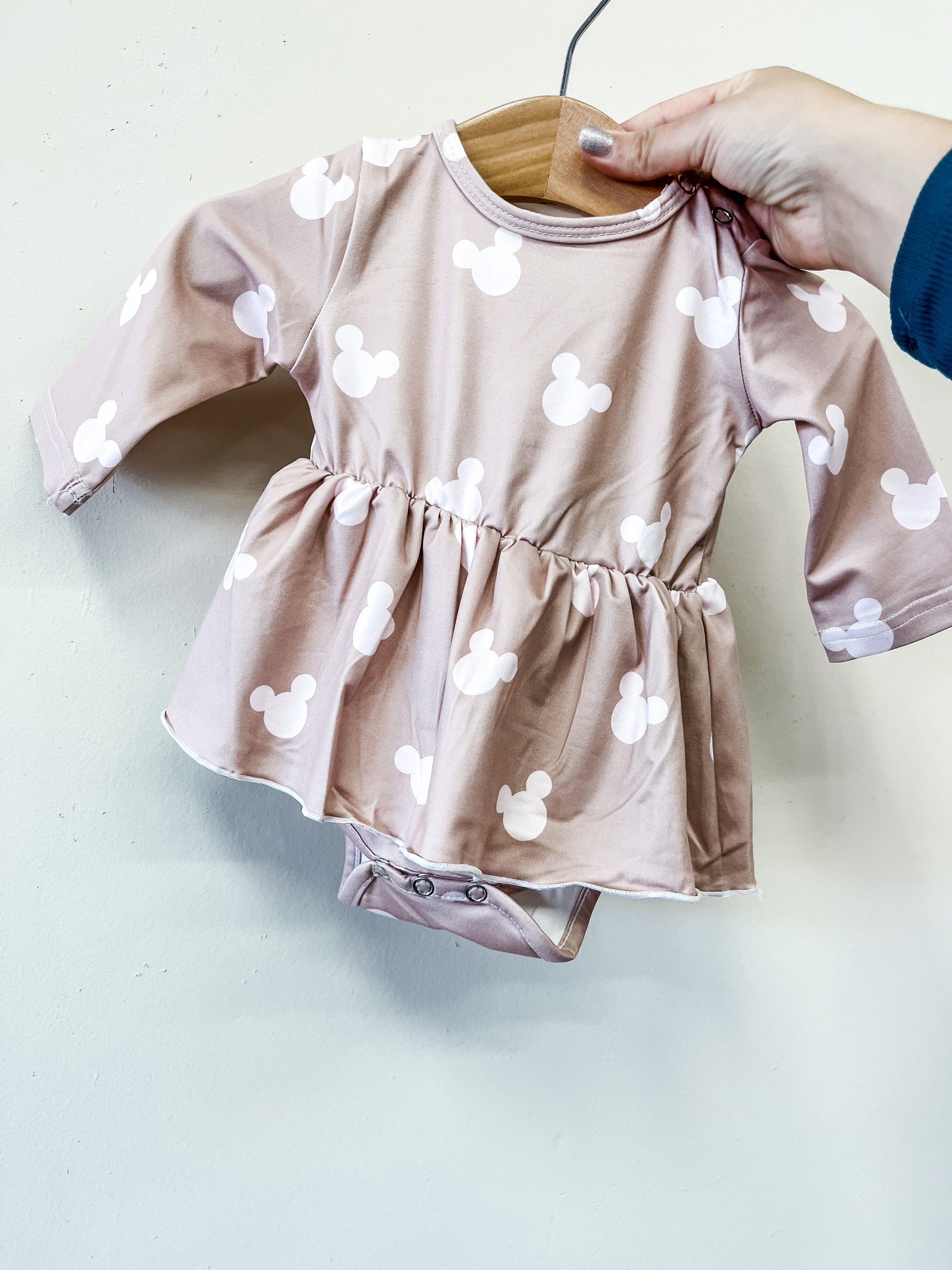 A hand displays the Cozy Mouse Longsleeve Twirl Romper by Tiny Tot Bundles—a pink baby romper with long sleeves, flared skirt, and white Mickey Mouse head prints—on a wooden hanger against a light background.
