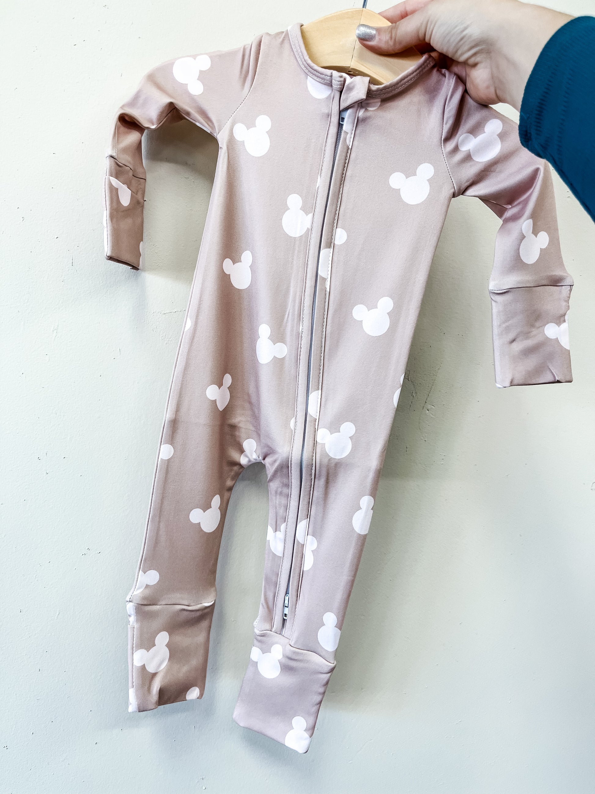 A hand holds up the Cozy Mouse Zippy by Tiny Tot Bundles, a mauve baby onesie with a front zipper and white Mickey Mouse head prints, against a plain light background.