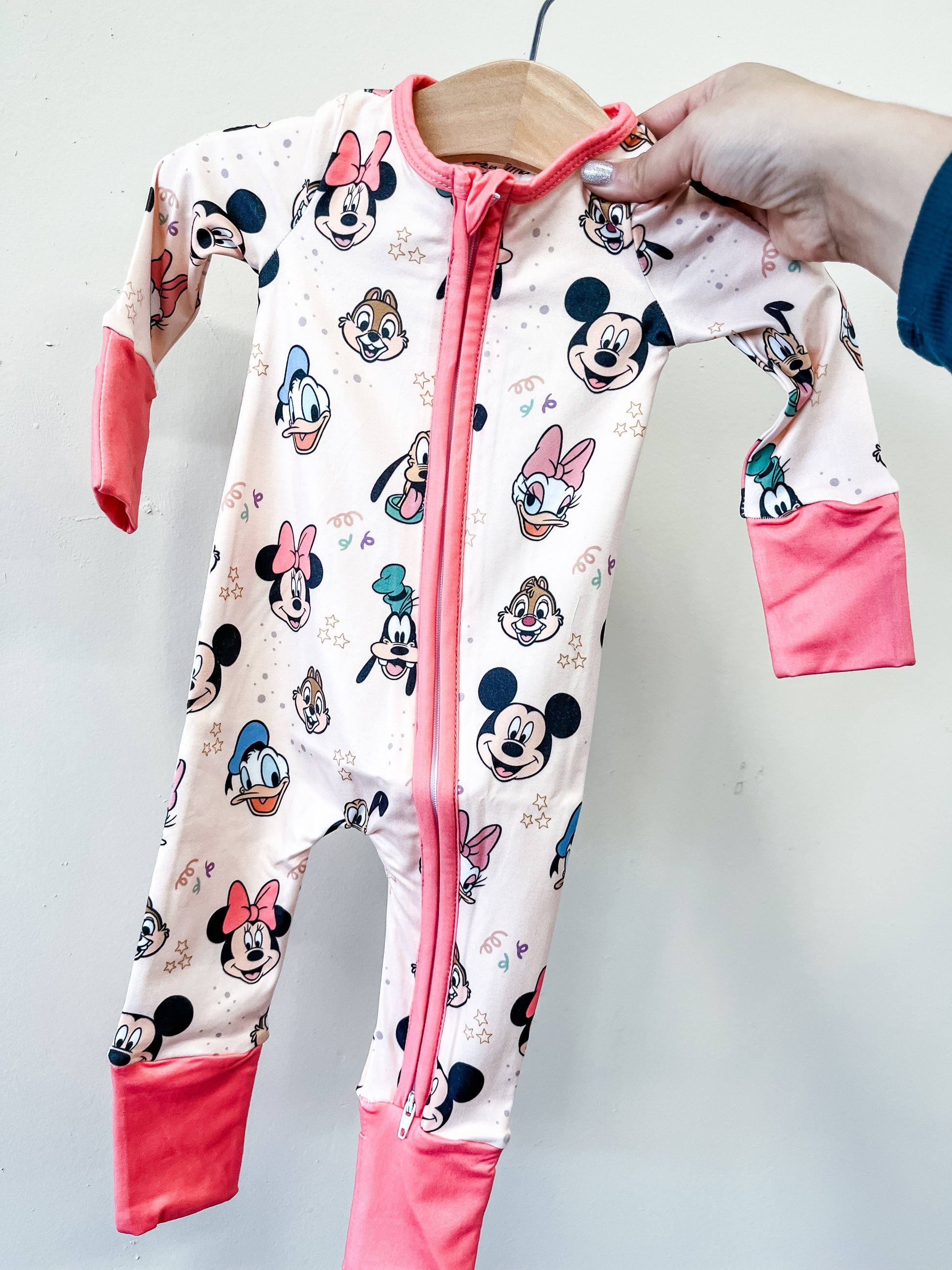 A hand holds up the Tiny Tot Bundles Magic Friends Zippy, a baby onesie with a front zipper, featuring colorful Disney character faces—Mickey Mouse, Minnie Mouse, Donald Duck, and Daisy Duck—with pink cuffs and trim.