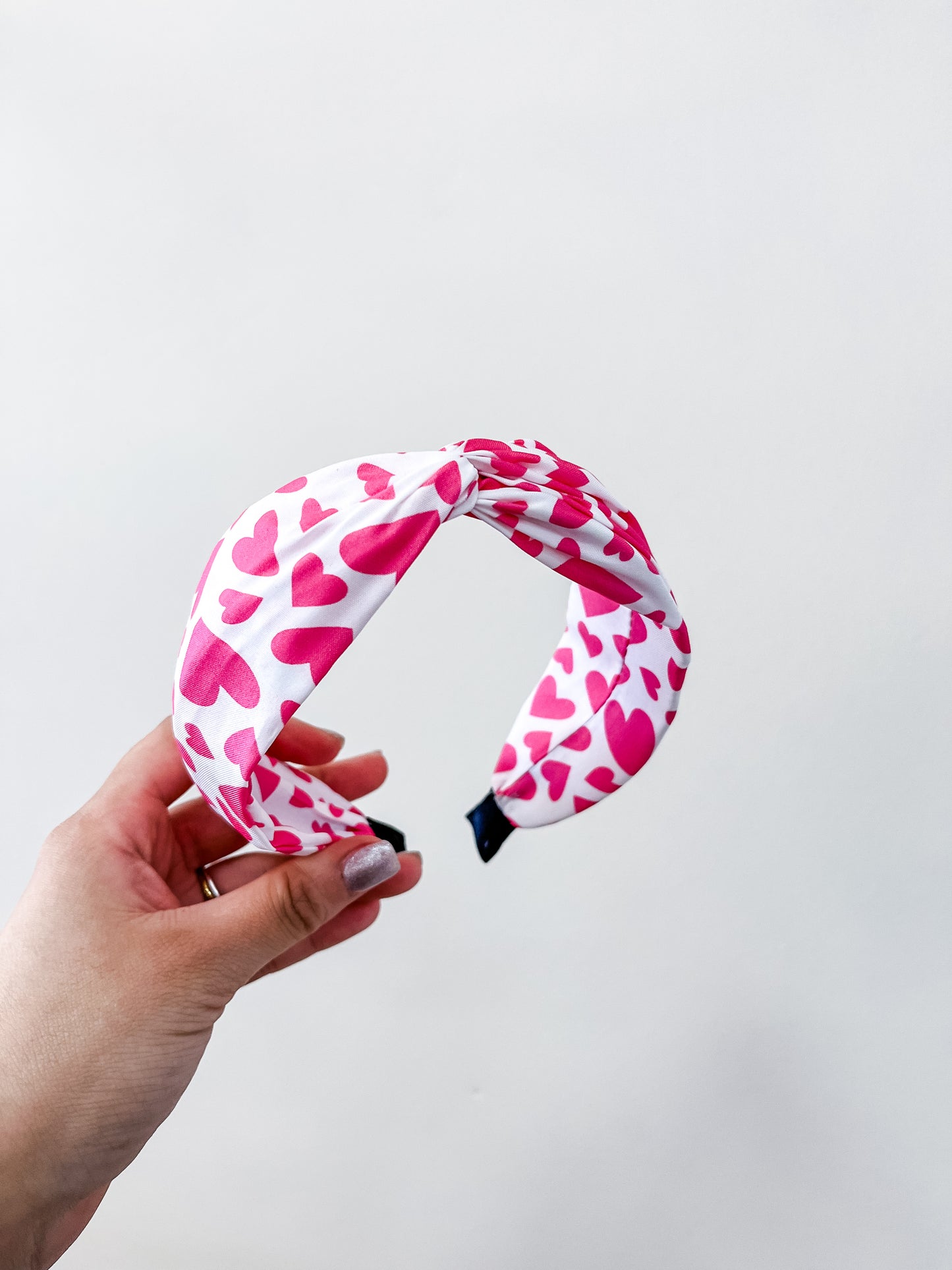 A hand holds the love & repeat Sweetheart Knot Headband, a white accessory featuring a twist design with pink hearts in various sizes, set against a plain white background.
