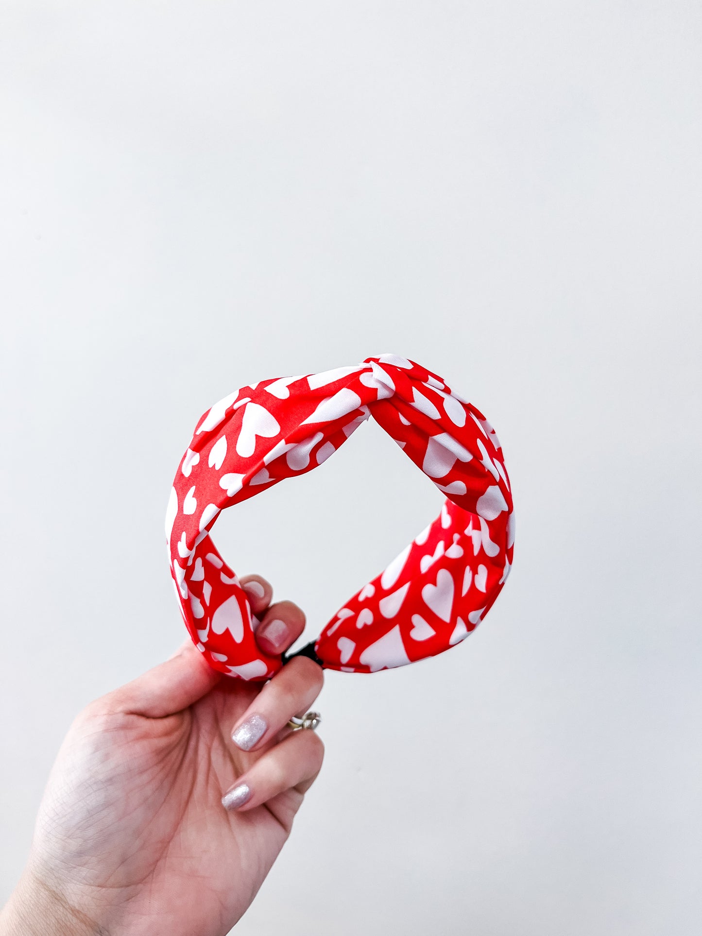 A hand holds the Sweetheart Knot Headband by love & repeat, featuring a red band adorned with white hearts, set against a plain white background.