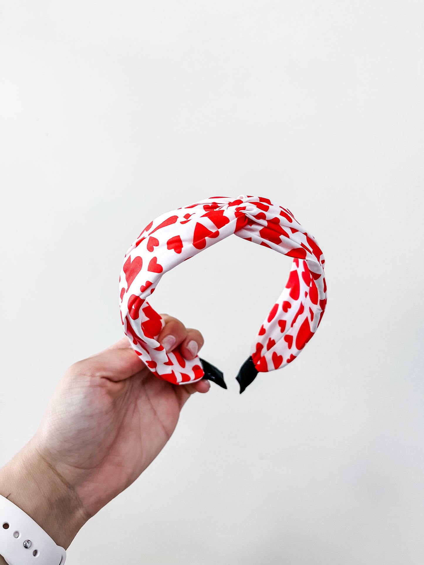 A hand holds the love & repeat Sweetheart Knot Headband, a white accessory adorned with red heart patterns, against a plain white background.