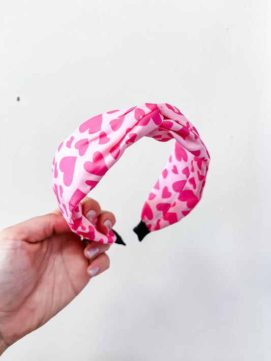 A hand holds the love & repeat Sweetheart Knot Headband, featuring pink fabric adorned with darker pink hearts, set against a plain white background.