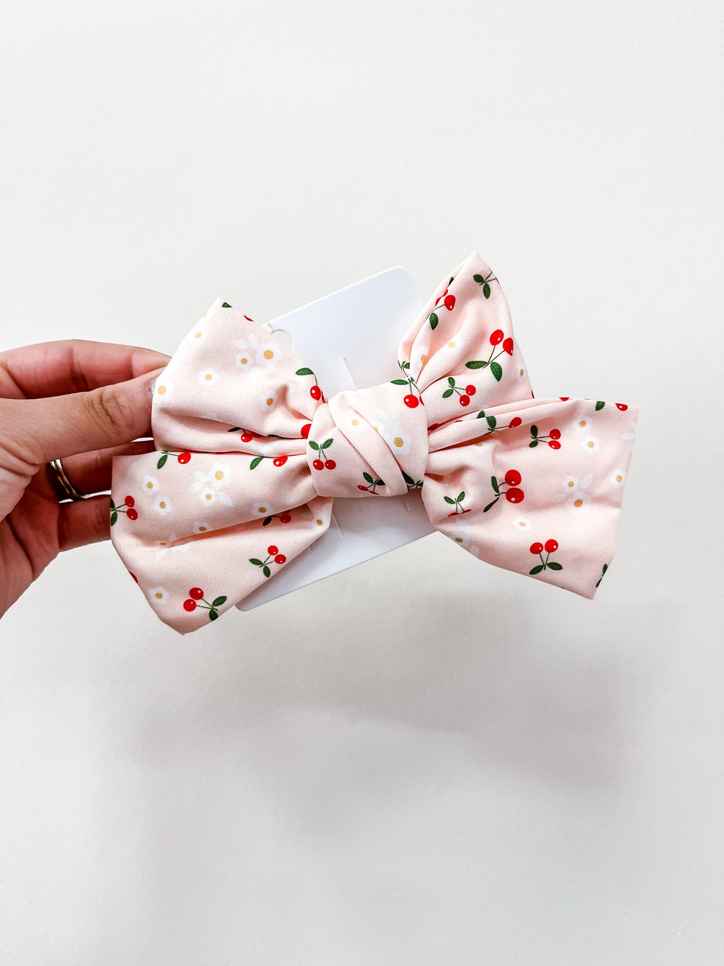 A hand holds a love & repeat Sweet Cherry Days Hair Bow Clip, featuring small red cherries and white flowers, against a plain white background.