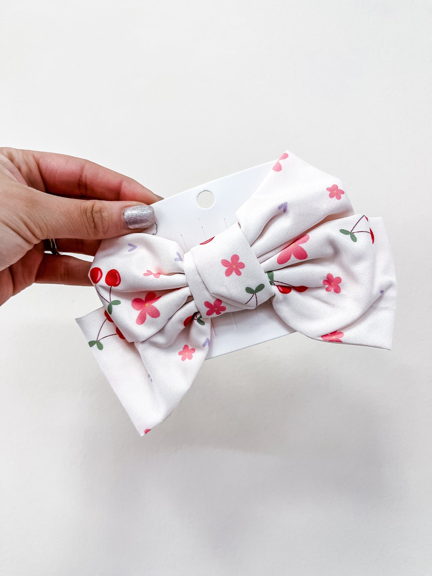 A hand with metallic nail polish holds the love & repeat Sweet Cherry Days Hair Bow Clips, featuring pink flowers and red cherries on a white bow, attached to a display card against a plain background.