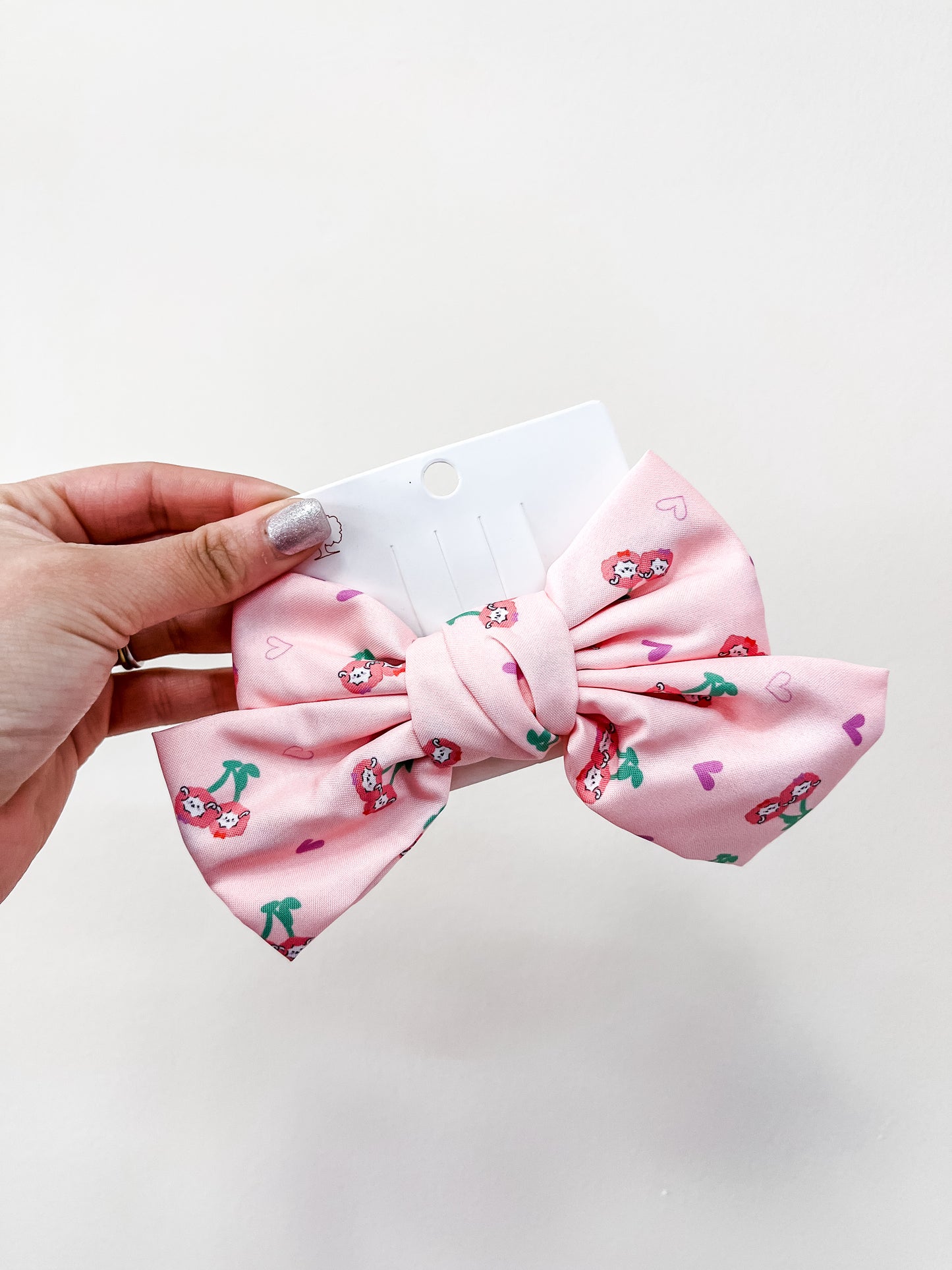 A hand holds a large pink Sweet Cherry Days Hair Bow Clip by love & repeat, featuring strawberry and heart patterns, displayed on a white card against a plain light background.
