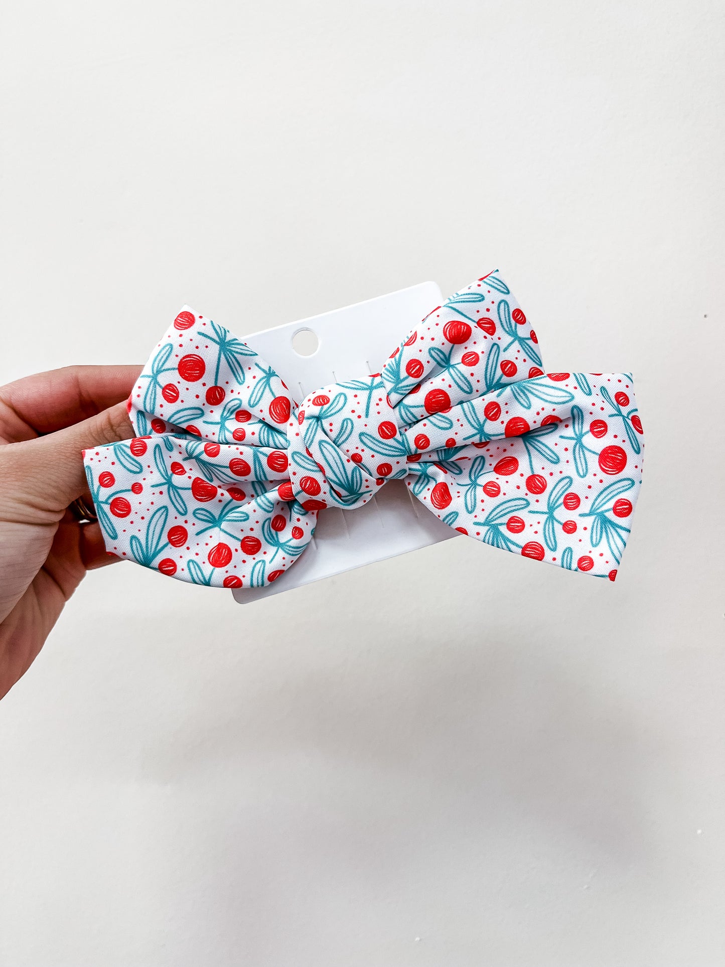 A hand holds a love & repeat Sweet Cherry Days Hair Bow Clip—large, white, with red cherries and green leaves—attached to a white card against a plain background.