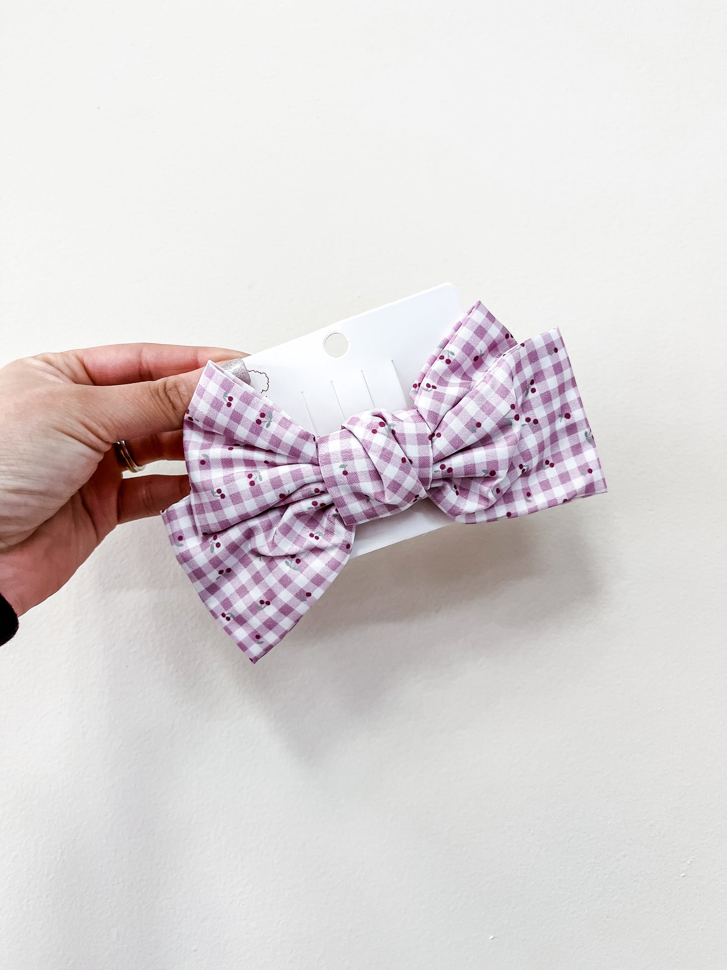 A hand holds a white card featuring a large Sweet Cherry Days Hair Bow Clip by love & repeat, showcasing a pink and white gingham pattern with small dotted accents against a plain white background.