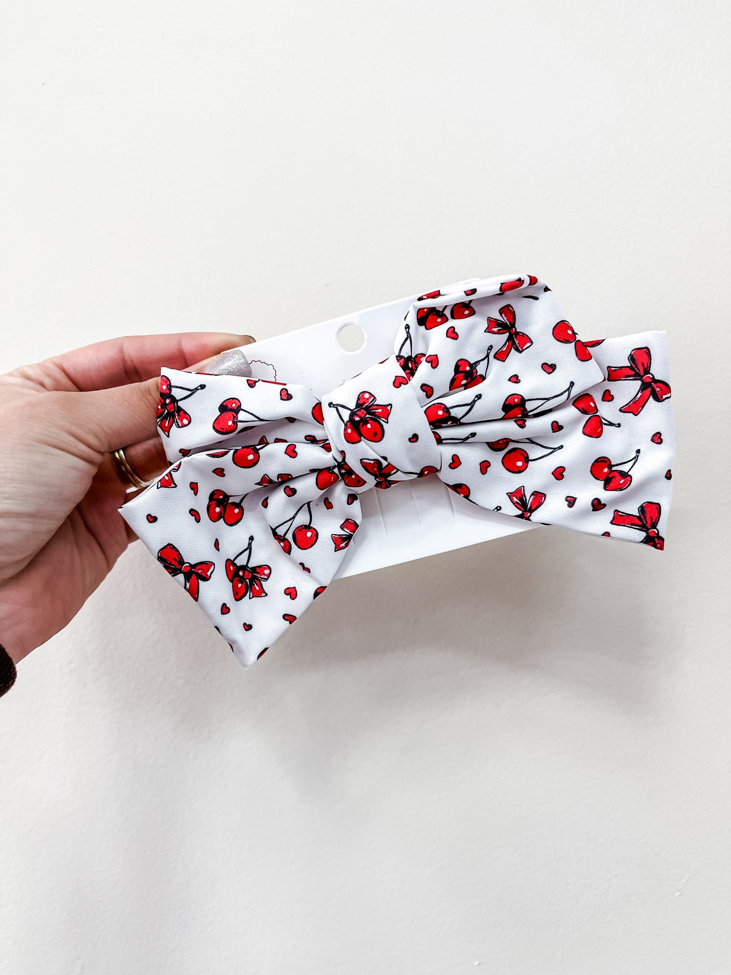 A hand holds a love & repeat Sweet Cherry Days Hair Bow Clip, featuring a large white bow with small red cherries and bows, against a plain white background.