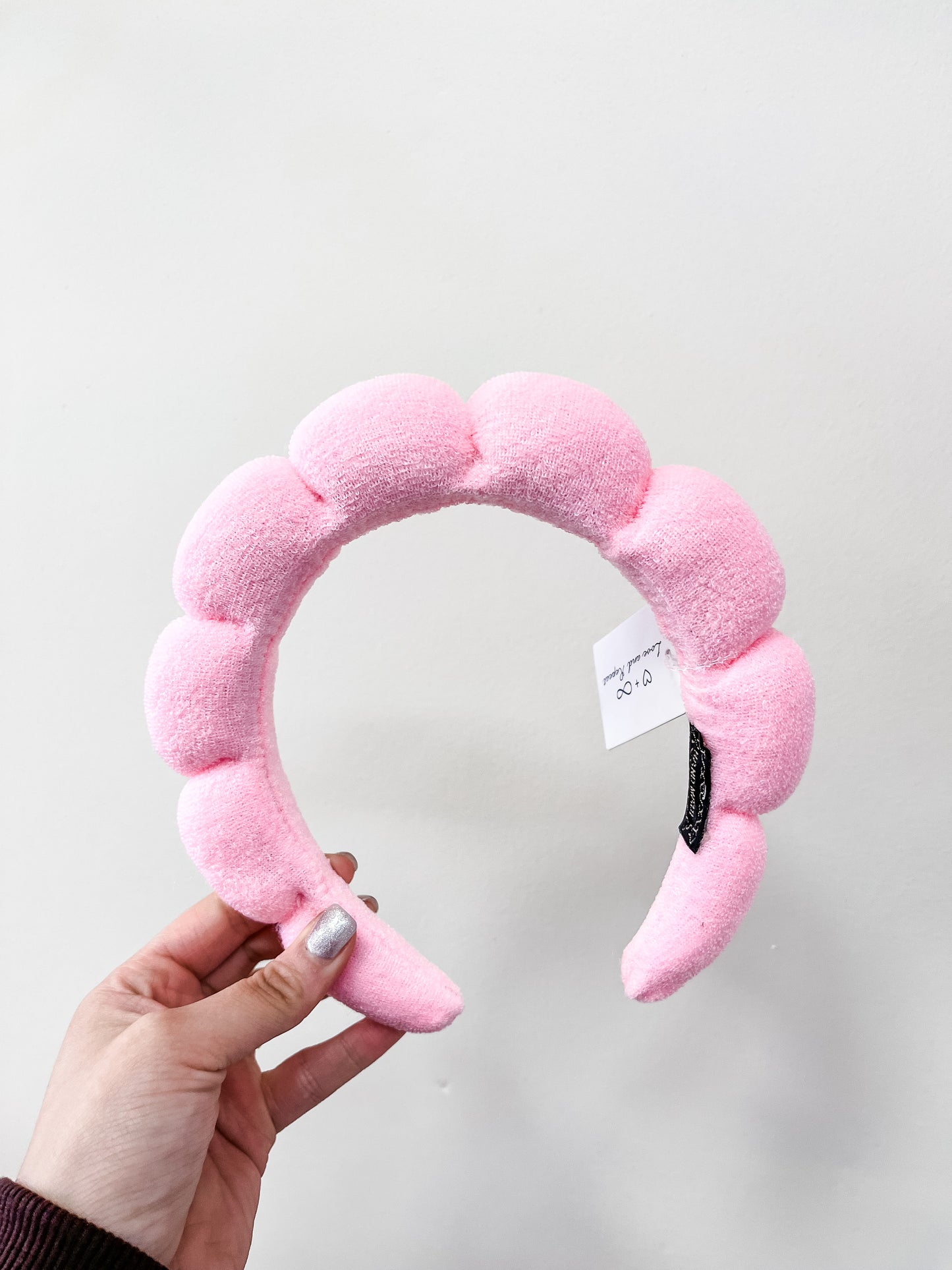 A hand holds the love & repeat Cloud Puff Headband, a soft, textured light pink accessory with a white price tag, set against a plain white background.