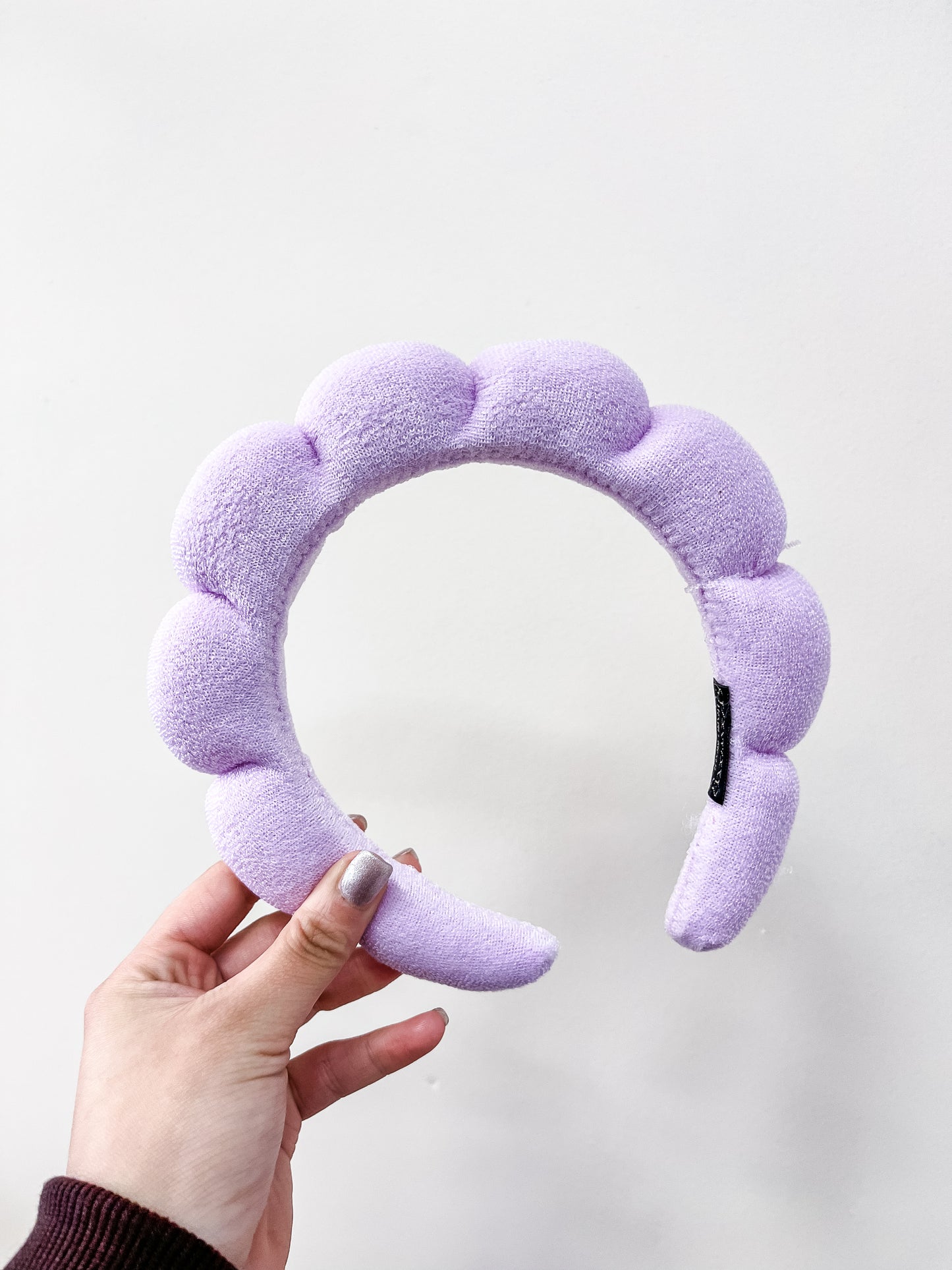 A hand with painted nails holds the love & repeat Cloud Puff Headband, a light purple plush headband with a scalloped design, against a plain white background.