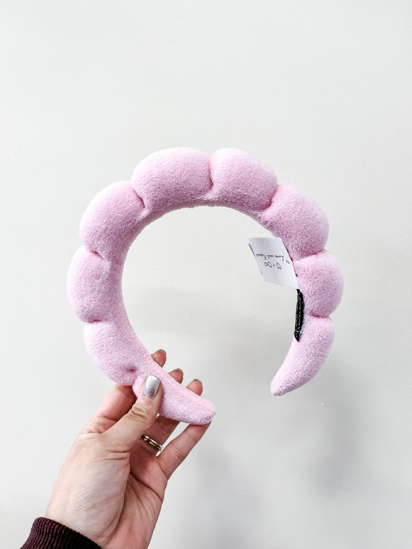 A hand holds the Cloud Puff Headband by love & repeat—light pink, padded fabric with a scalloped design and a small white tag—set against a plain white background.