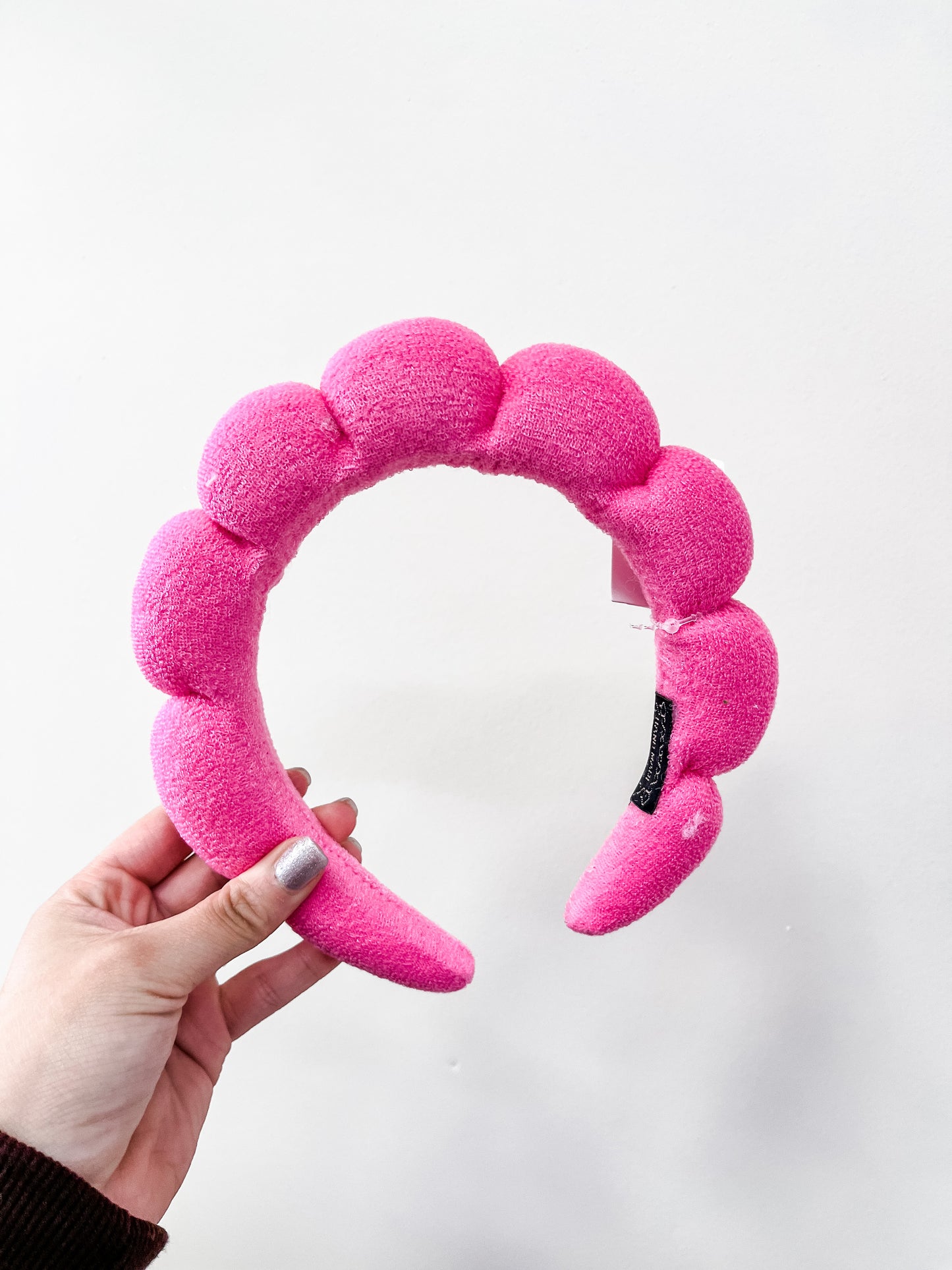 A hand with silver nail polish holds the bright pink Cloud Puff Headband by love & repeat, featuring a plush, bubble-shaped design, against a plain white background.
