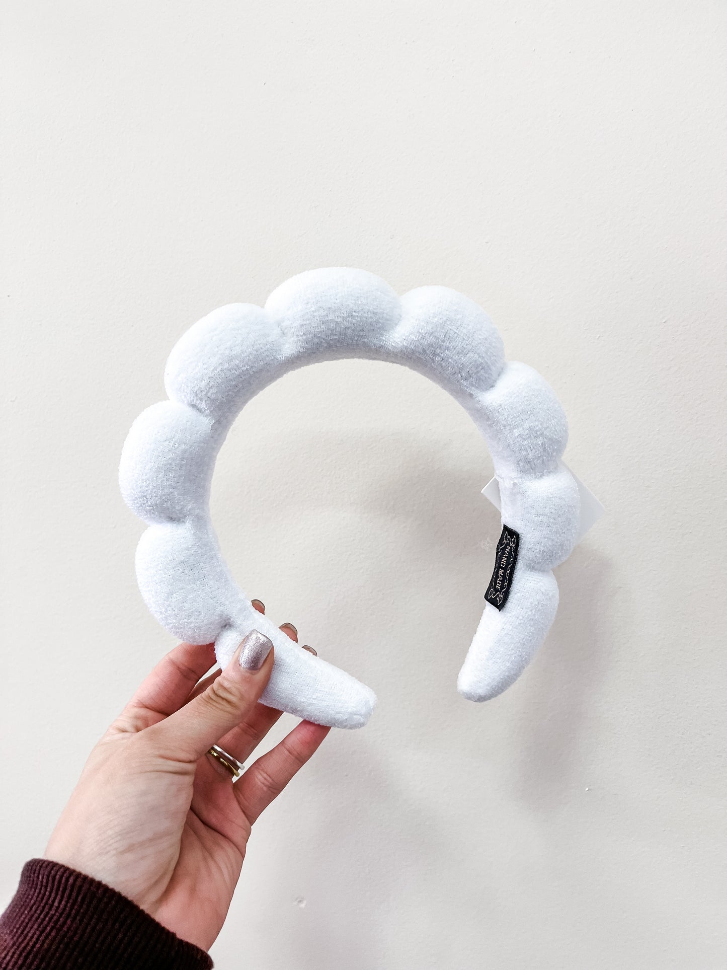 A hand with painted nails holds the love & repeat Cloud Puff Headband, a plush white scalloped accessory, against a plain off-white background.