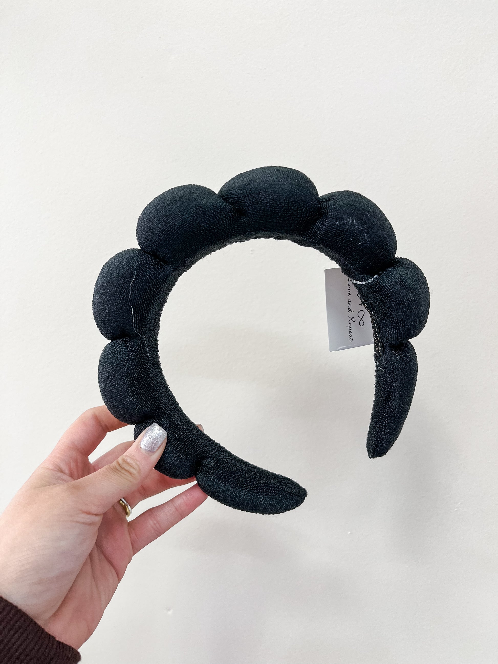 A hand with metallic silver nail polish holds the love & repeat Cloud Puff Headband—black, padded, and scalloped—against a plain white background. A small tag is attached to the headband.