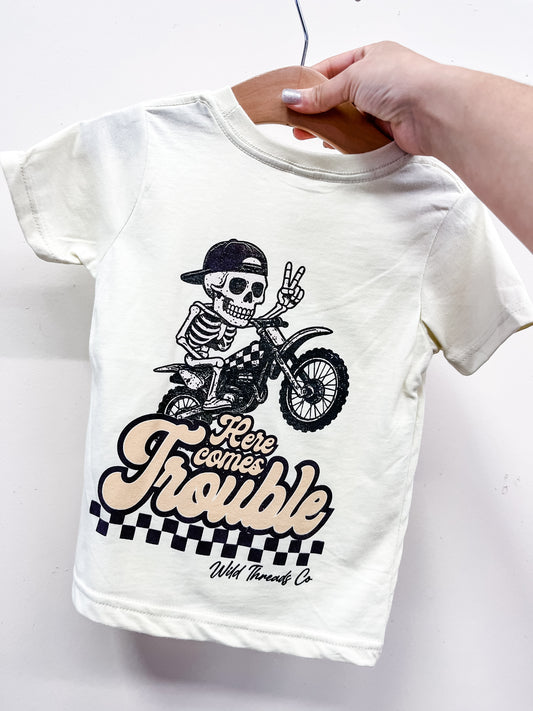 A hand holds the "Here Comes Trouble" T-shirt by Wild Threads Co, featuring a skeleton on a motorcycle in a backwards cap giving a peace sign, checkered pattern accents, and playful text.