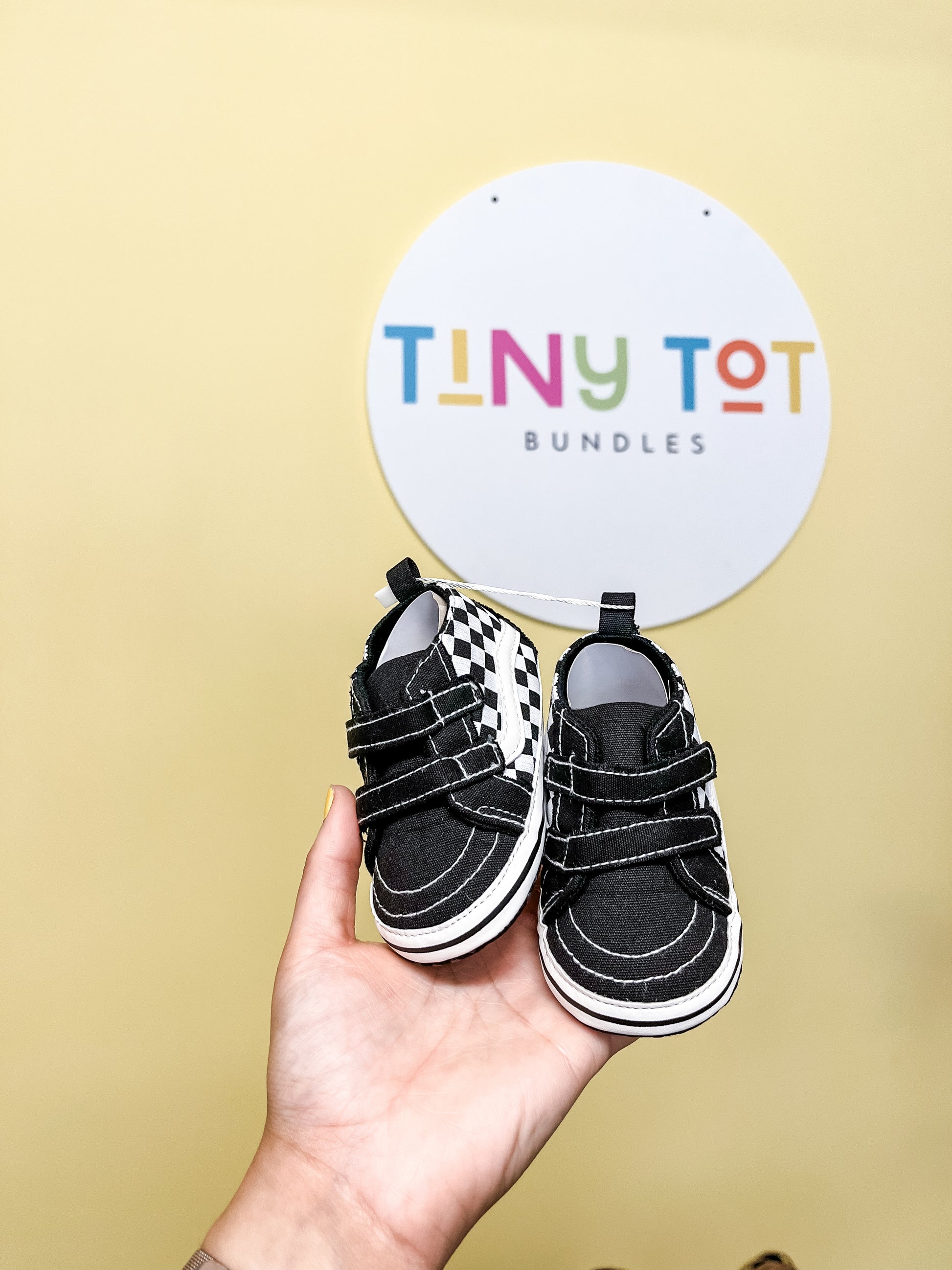 A hand holds up a pair of Tiny Tot Bundles Checkered Hightops—small black and white baby shoes—in front of a colorful "Tiny Tot Bundles" wall sign.