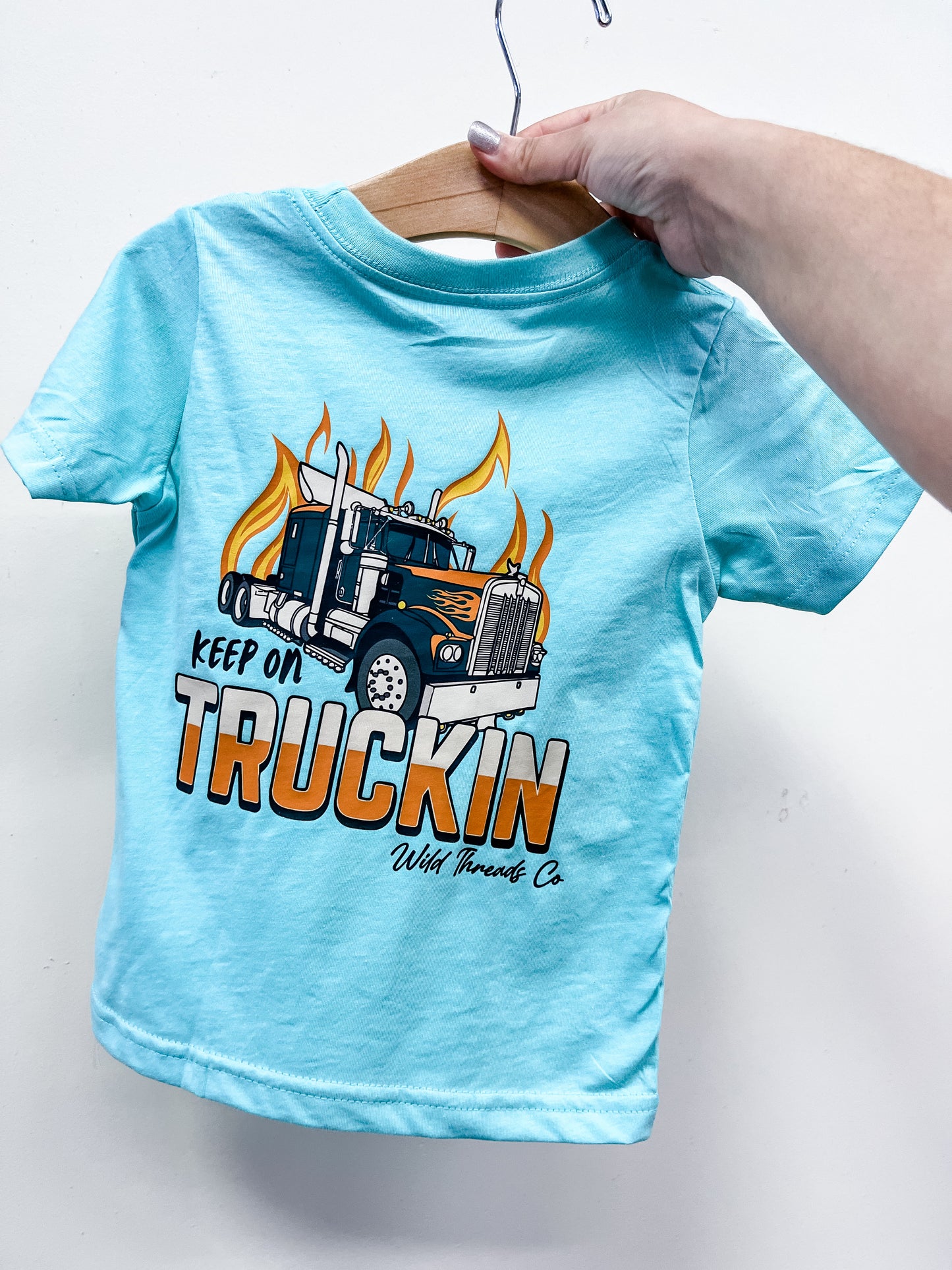 A hand holds a light blue Keep On Truckin' T-Shirt by Wild Threads Co on a wooden hanger, featuring a bold graphic of a semi-truck with flames and the words “Keep On Truckin’”.