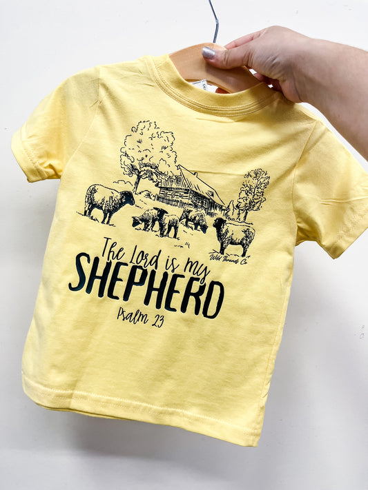 A hand holds Wild Threads Co’s "The Lord Is My Shepperd" T-shirt in yellow, featuring black sheep, trees, a barn, and the text "The Lord is my Shepherd, Psalm 23" on the front.