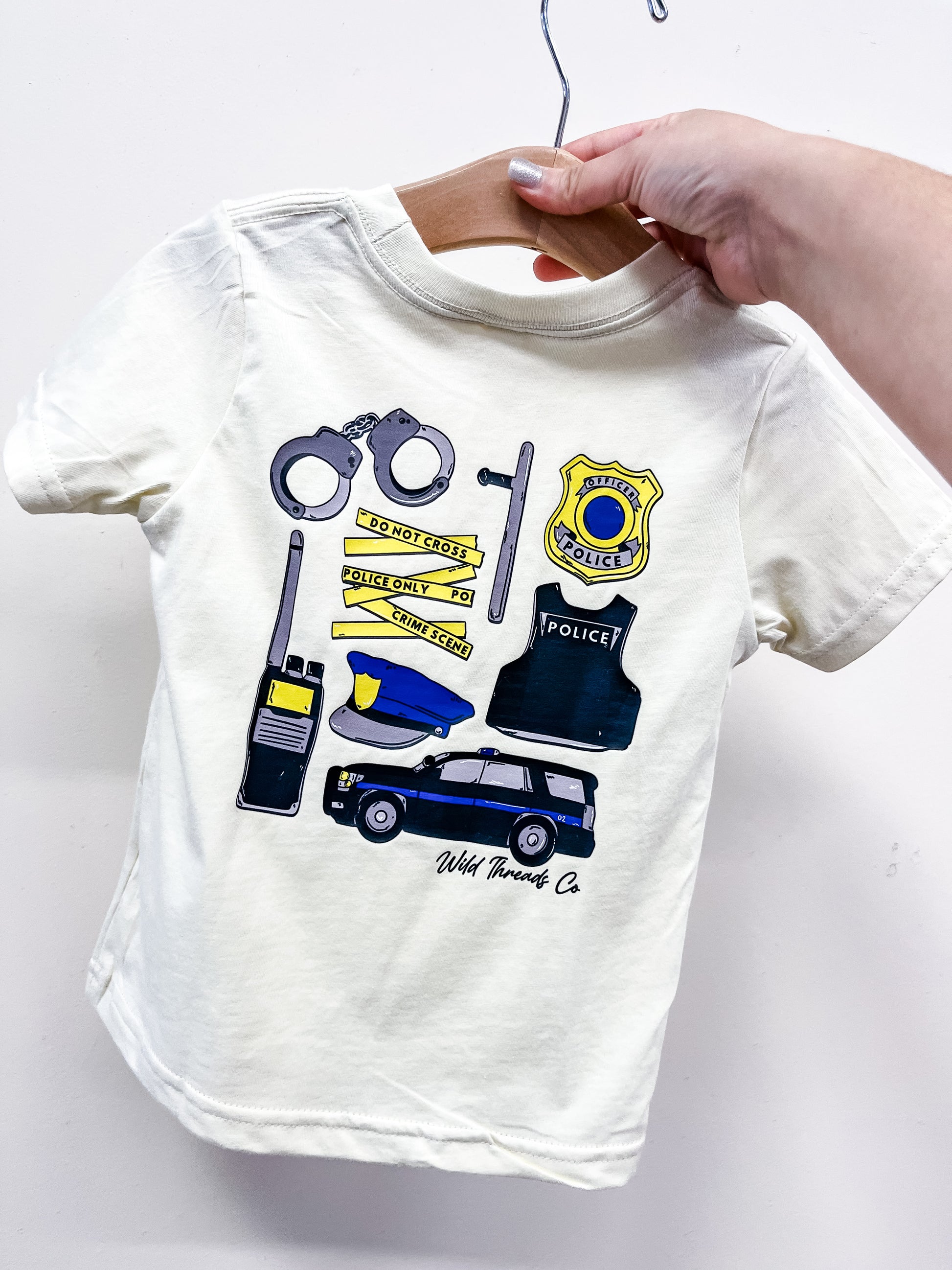A hand holds a white toddler "Tool Of The Trade - Police Officer T-Shirt" by Wild Threads Co on a wooden hanger, featuring police-themed illustrations like handcuffs, badge, hat, tape, radio, patrol car, and the brand name.