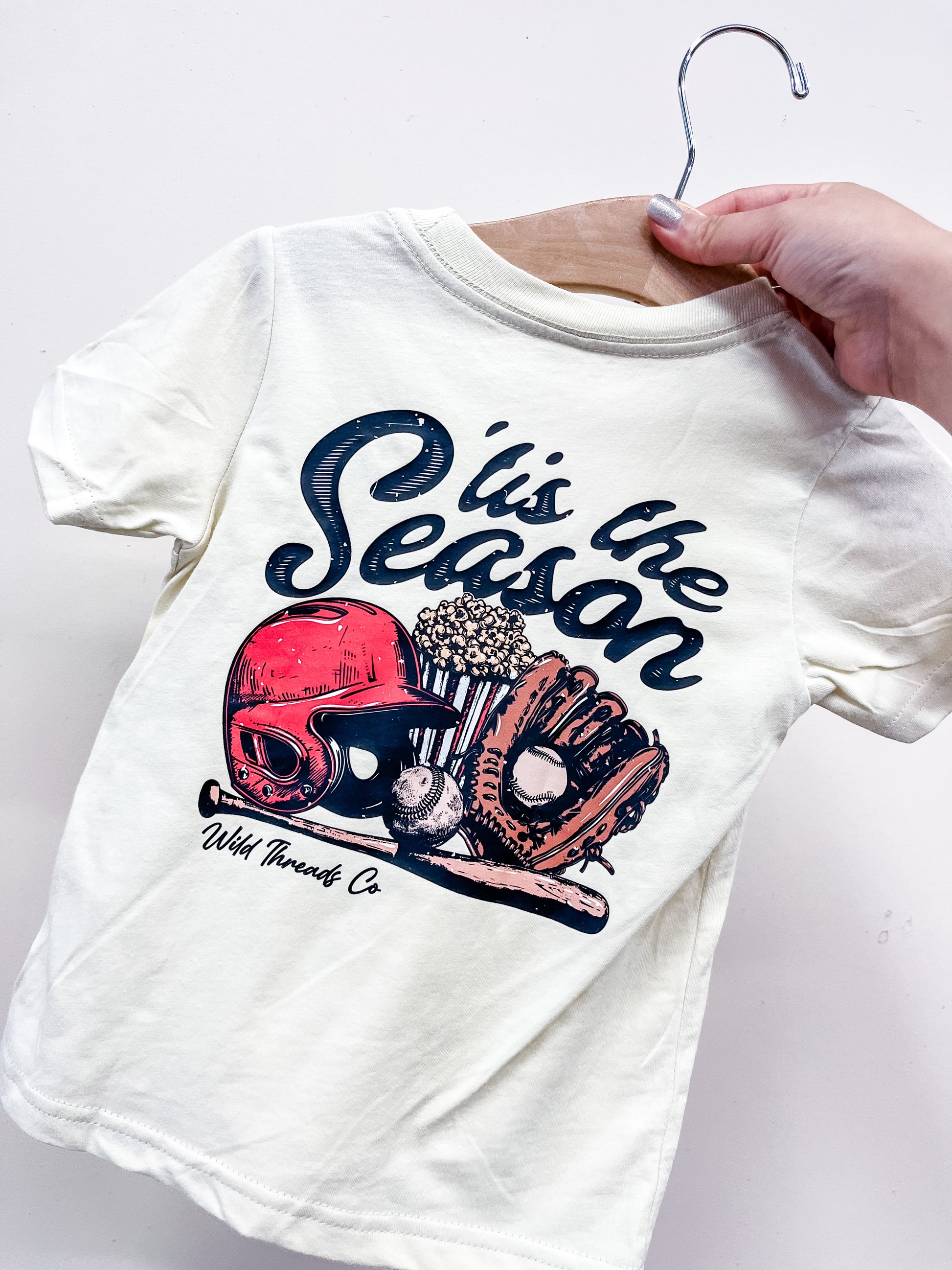 A hand holds the Tis' The Season Baseball T-Shirt by Wild Threads Co, featuring a red baseball helmet, glove, ball, bats, and the text "tis the Season" in a stylized font.