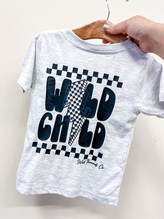 A hand holds the Wild Threads Co Wild Child Lightning Bolt T-Shirt on a wooden hanger. The back features bold blue "WILD CHILD" text, a checkered lightning bolt, and checkerboard accents.