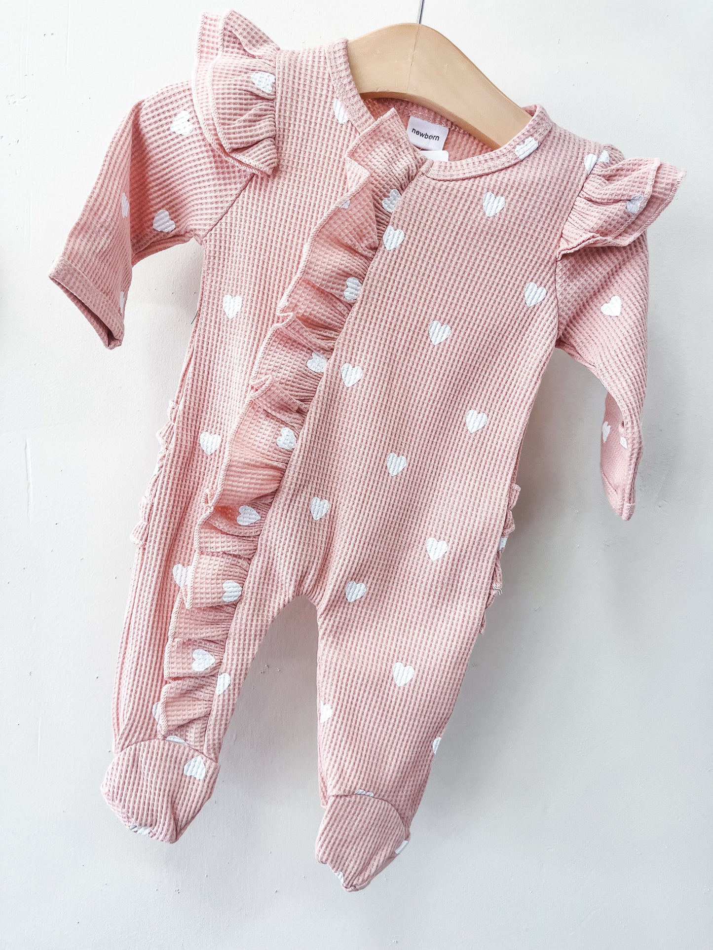 The Little Sweetheart Waffle Footie by Tiny Tot Bundles is a pink long-sleeve baby onesie with white heart patterns, ruffled trim on the front and shoulders, and footed legs, shown on a wooden hanger against a white background.