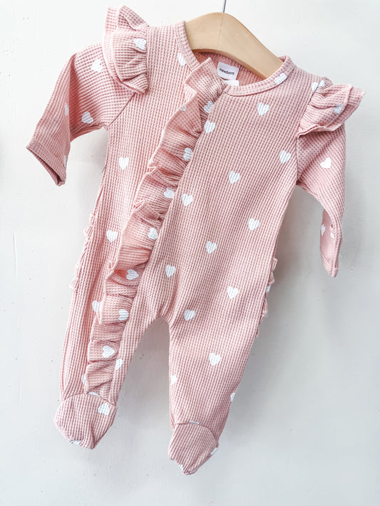 The Little Sweetheart Waffle Footie by Tiny Tot Bundles is a pink long-sleeve baby onesie with white heart patterns, ruffled trim on the front and shoulders, and footed legs, shown on a wooden hanger against a white background.