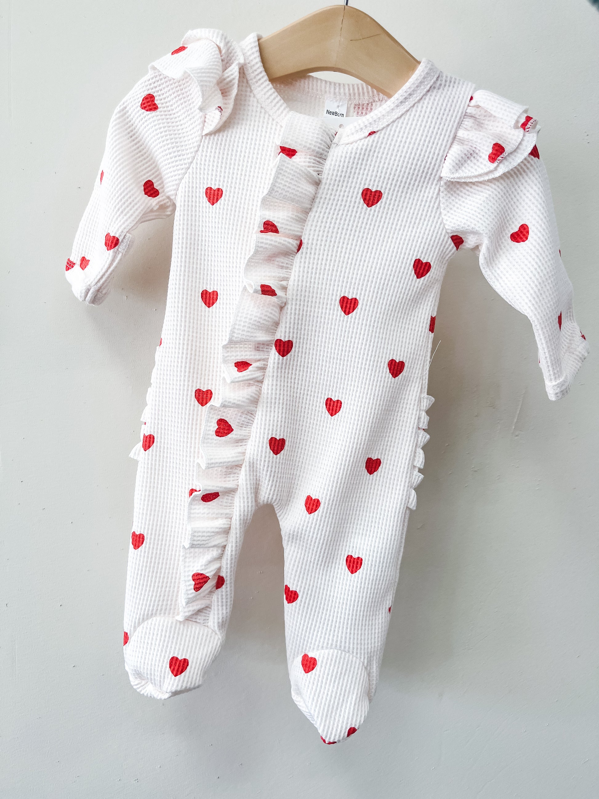 The Little Sweetheart Waffle Footie by Tiny Tot Bundles is a white baby onesie with red heart prints, ruffled details on the front and shoulders, and covered feet, shown hanging on a wooden hanger against a plain white background.