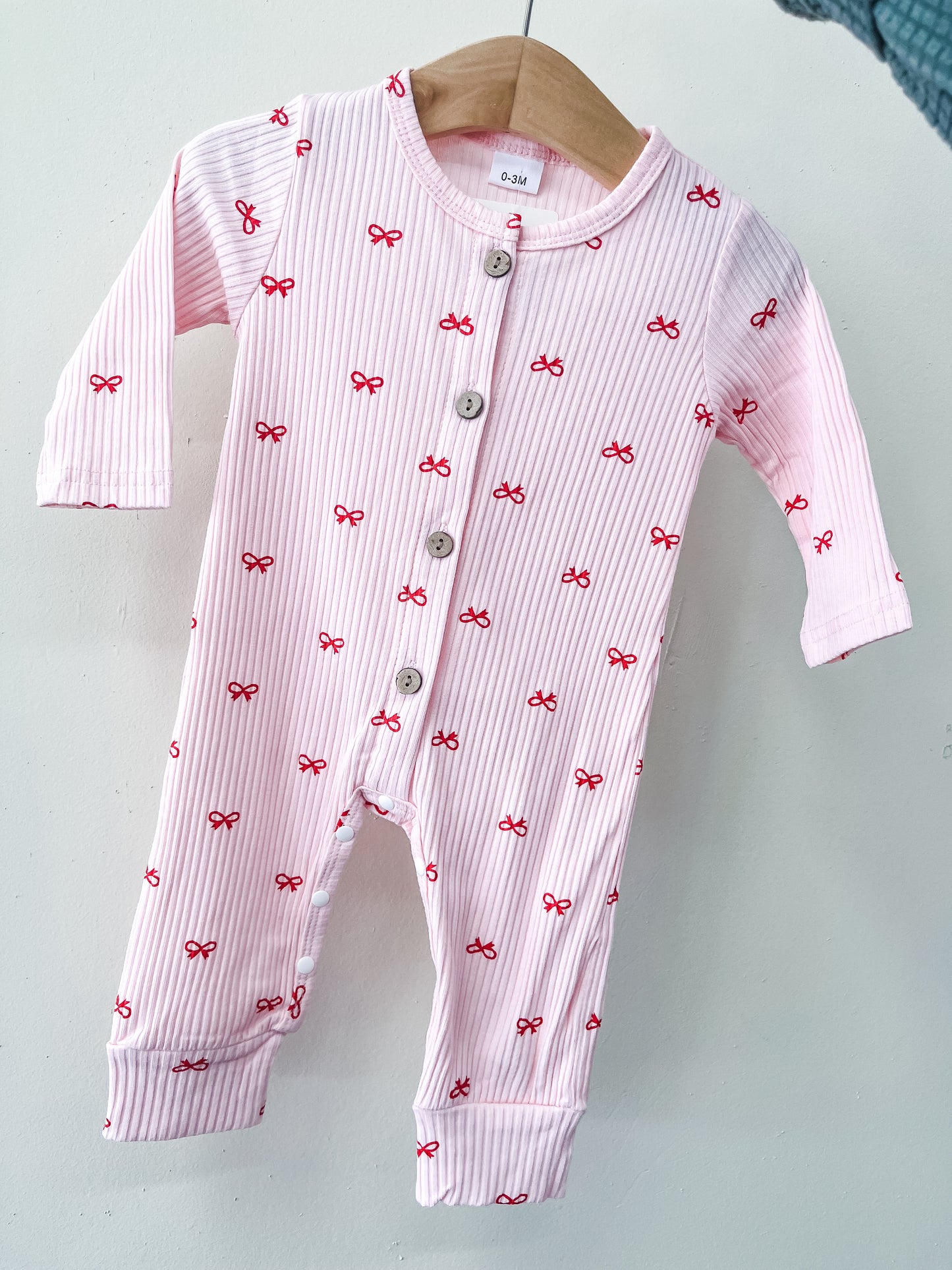 The Tiny Tot Bundles Tiny Bows Ribbed Romper, a pink, long-sleeved baby outfit with red bow patterns and front buttons, hangs on a wooden hanger against a white background.