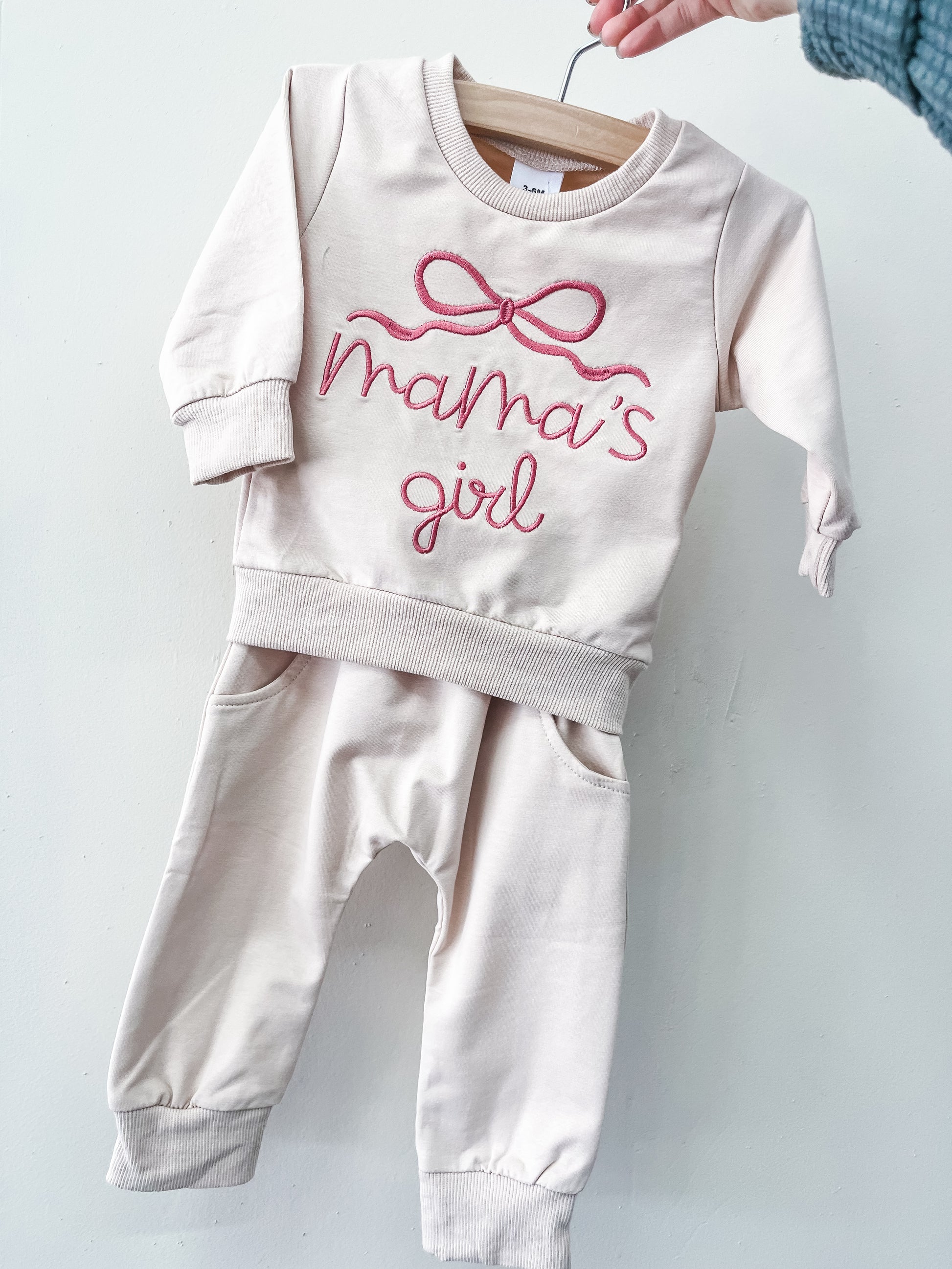 The Mama's Girl Cozy Set by Tiny Tot Bundles is a light pink baby outfit with long sleeves and pants, featuring "mamas girl" and a pink bow on the top, displayed on a wooden hanger against a white background.