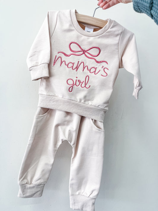 The Mama's Girl Cozy Set by Tiny Tot Bundles is a light pink baby outfit with long sleeves and pants, featuring "mamas girl" and a pink bow on the top, displayed on a wooden hanger against a white background.
