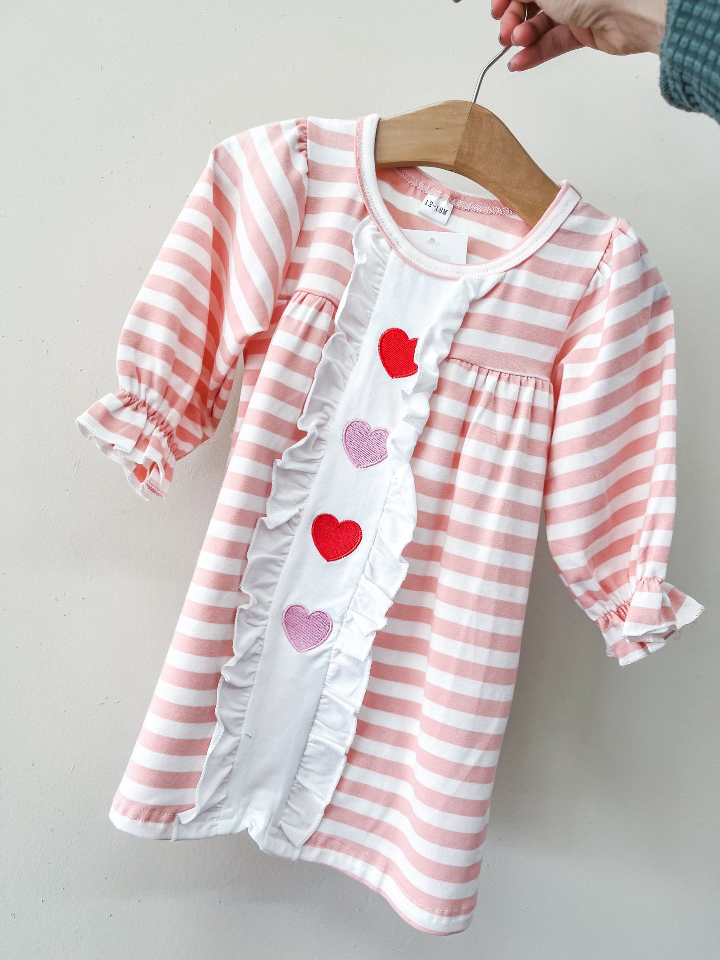 A hand displays the Rosy Ruffle Hearts Dress by Tiny Tot Bundles—a pink and white striped baby dress with ruffled sleeves, a white front ruffle panel, and red and pink heart appliqués.