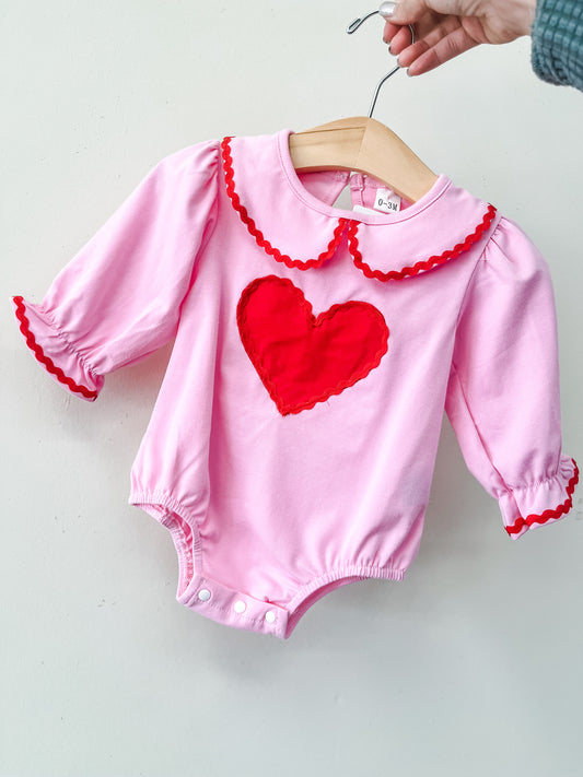 A hand holds the Tiny Tot Bundles Classic Sweetheart Romper, a pink long-sleeve onesie with red ruffled collar and cuffs, featuring a large red heart appliqué on the front.