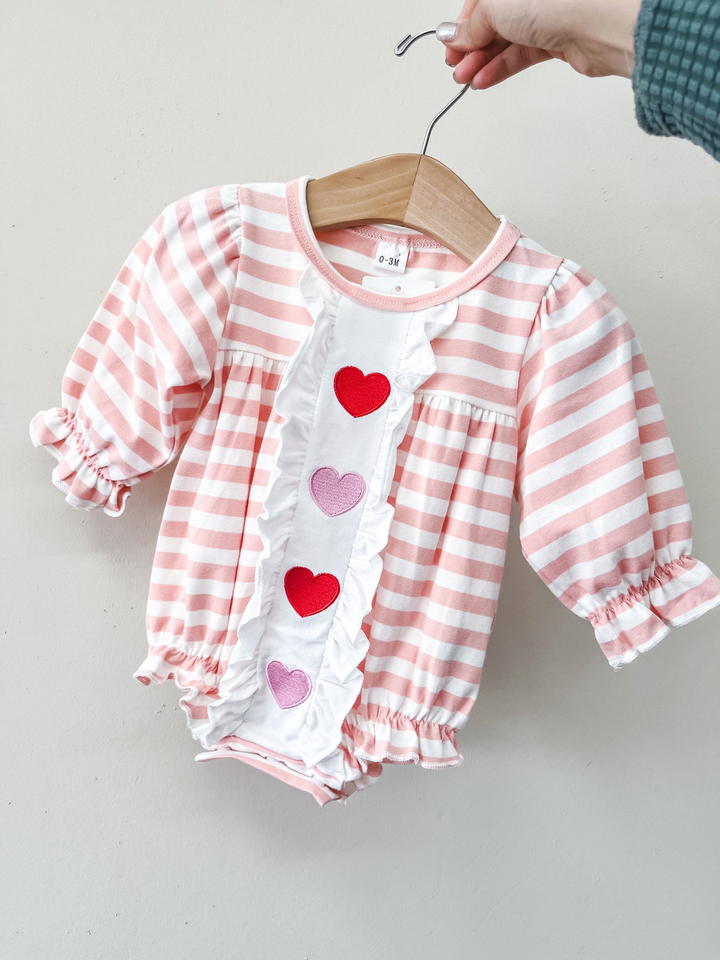 The Rosy Ruffles Hearts Romper by Tiny Tot Bundles features pink and white stripes, ruffled trim, and red and pink heart appliqués on the front. Shown on a hanger held by a hand.