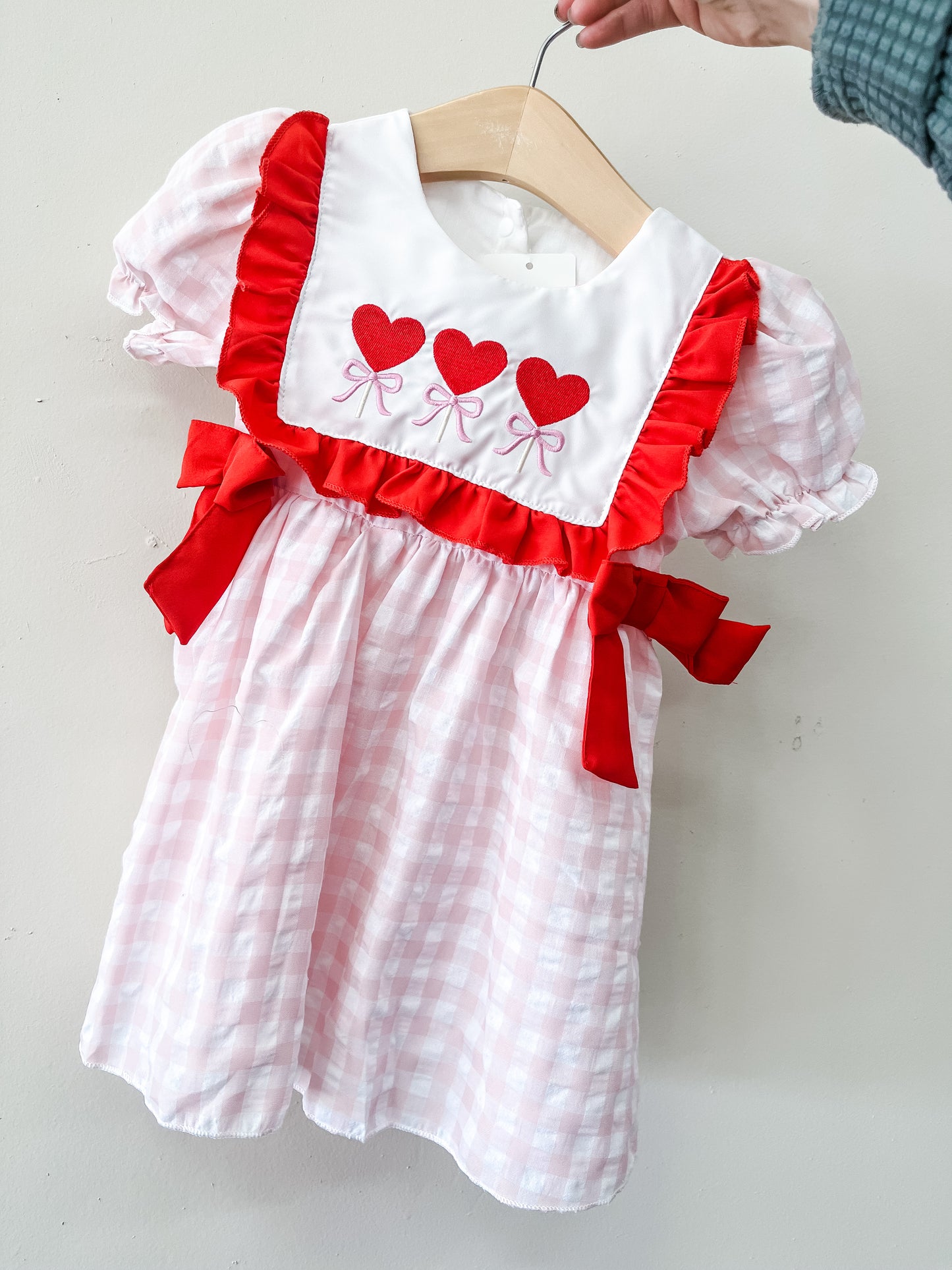 The Sweetheart Trio Dress by Tiny Tot Bundles is a pink and white checkered toddler dress with puffed sleeves, red ruffles and bows, plus a white bib adorned with three red hearts and pink bows, shown on a wooden hanger.