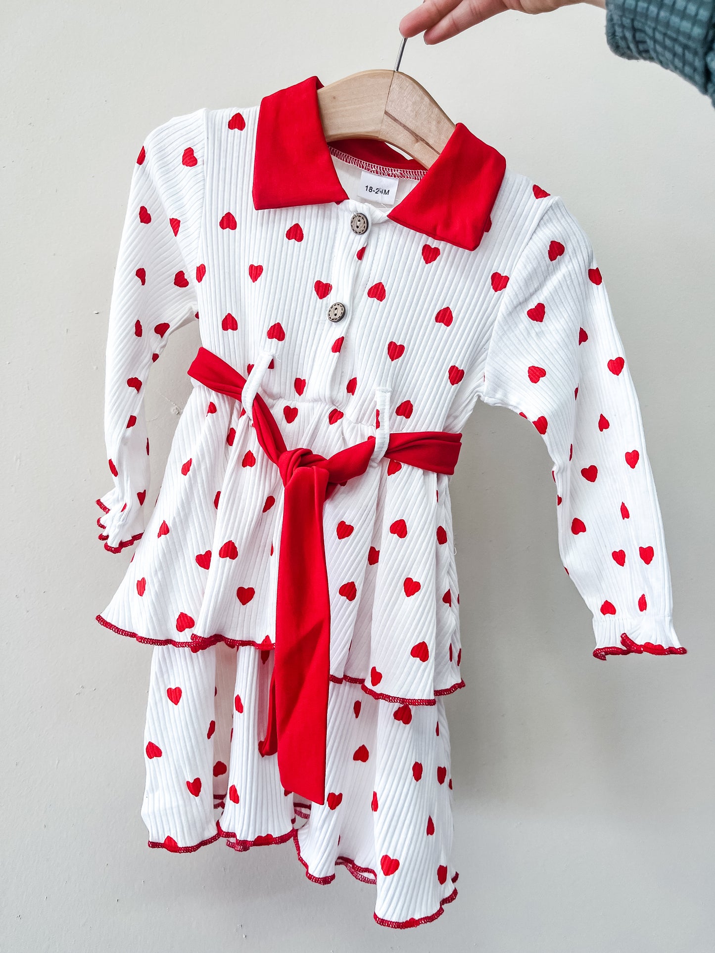 The Scarlet Hearts Tiered Dress by Tiny Tot Bundles features long sleeves, a white base with red heart prints, a red collar, matching waist belt, and a tiered skirt, displayed on a wooden hanger against a plain light background.