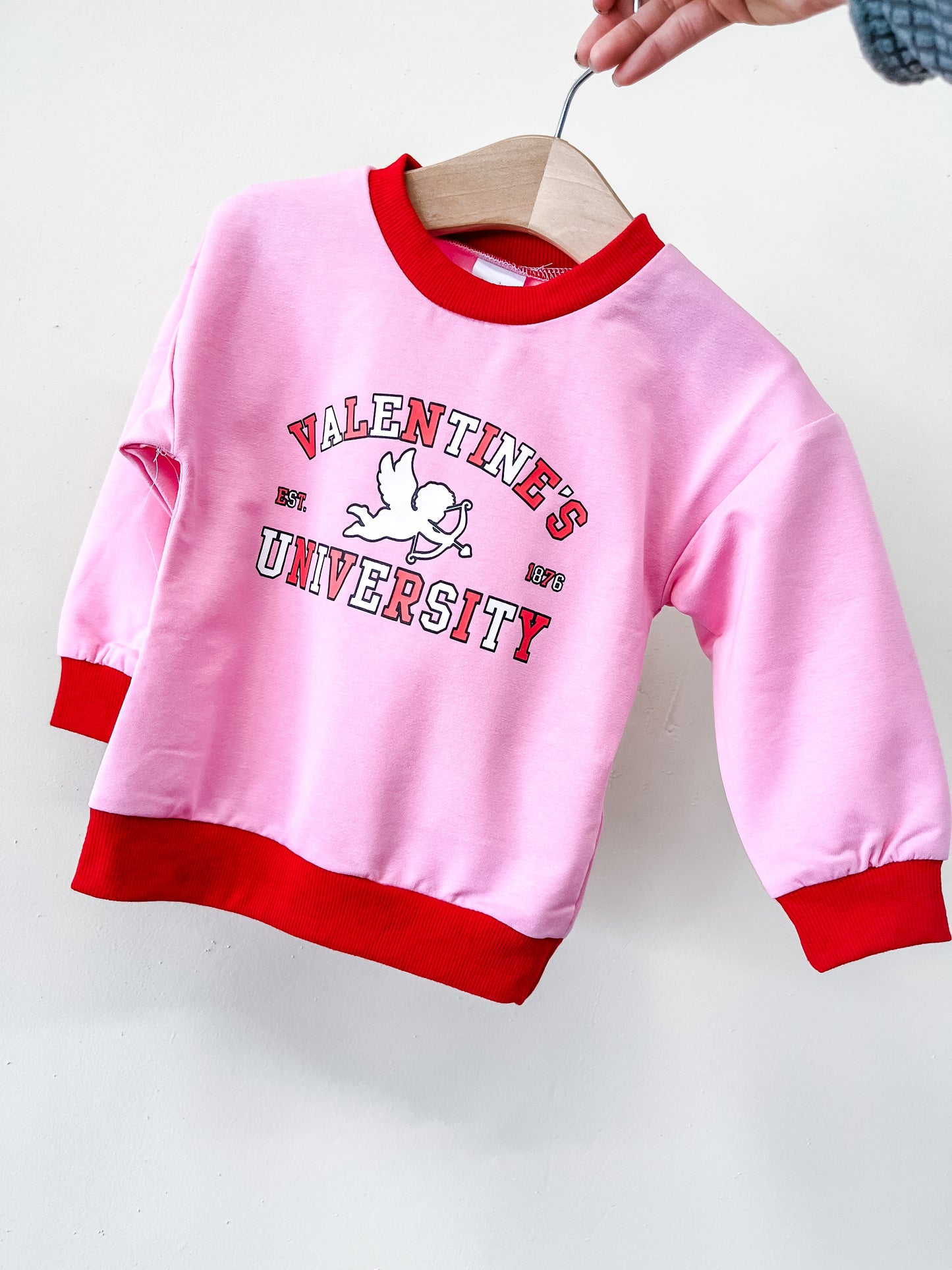 A hand holds up the Tiny Tot Bundles Valentines University Pullover—a pink sweatshirt with red cuffs and collar, featuring "Valentines University" text and a flying cupid—on a wooden hanger.