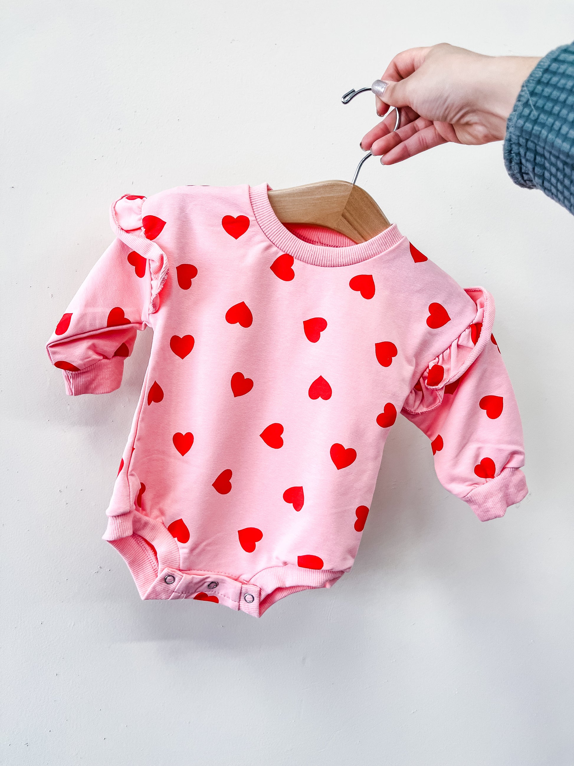 A hand holds a hanger with the Tiny Tot Bundles "All My Love Bubble," a pink long-sleeve baby onesie adorned with red heart patterns and ruffled shoulders, shown against a plain white background.