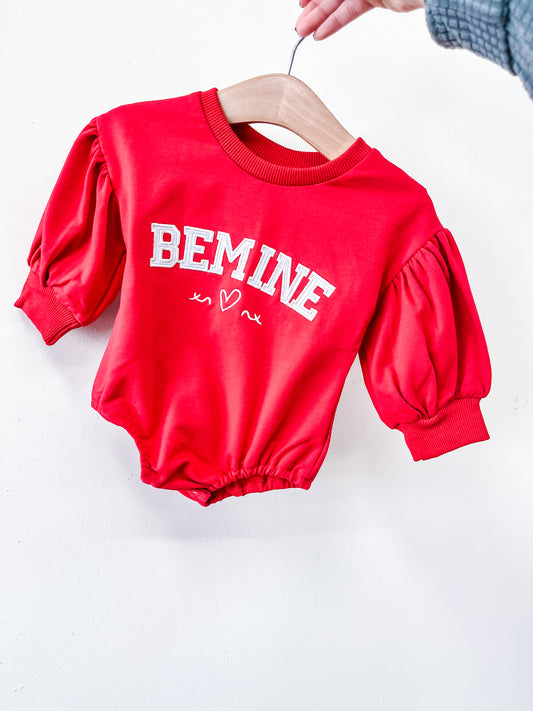 The Be Mine Puff Sleeve Bubble by Tiny Tot Bundles is a red baby romper with puffy sleeves, white "BE MINE" text, and two heart doodles on the front, displayed on a wooden hanger held by hand.