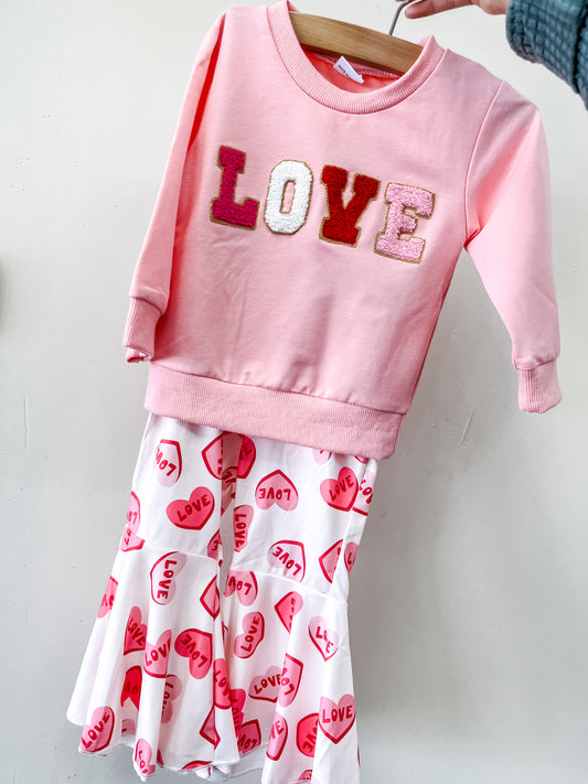 The Sweet Love Set by Tiny Tot Bundles features a pink LOVE-lettered sweatshirt and a white skirt with pink and red hearts plus repeated LOVE prints, displayed together on a hanger.